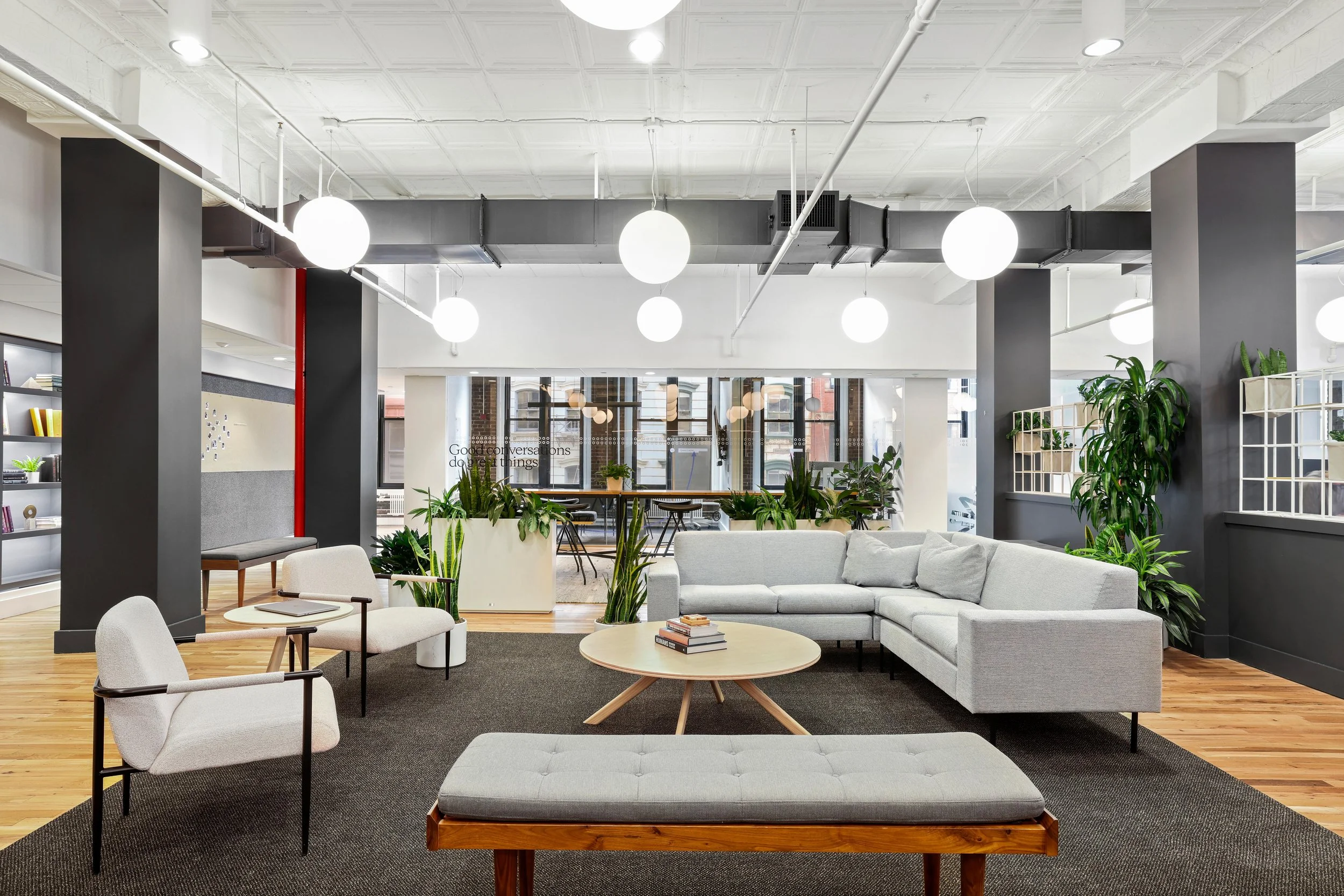 Modern office lounge with white sectional sofa, armchairs, round wooden coffee table with books, potted plants, black accent walls, and large windows letting in natural light.