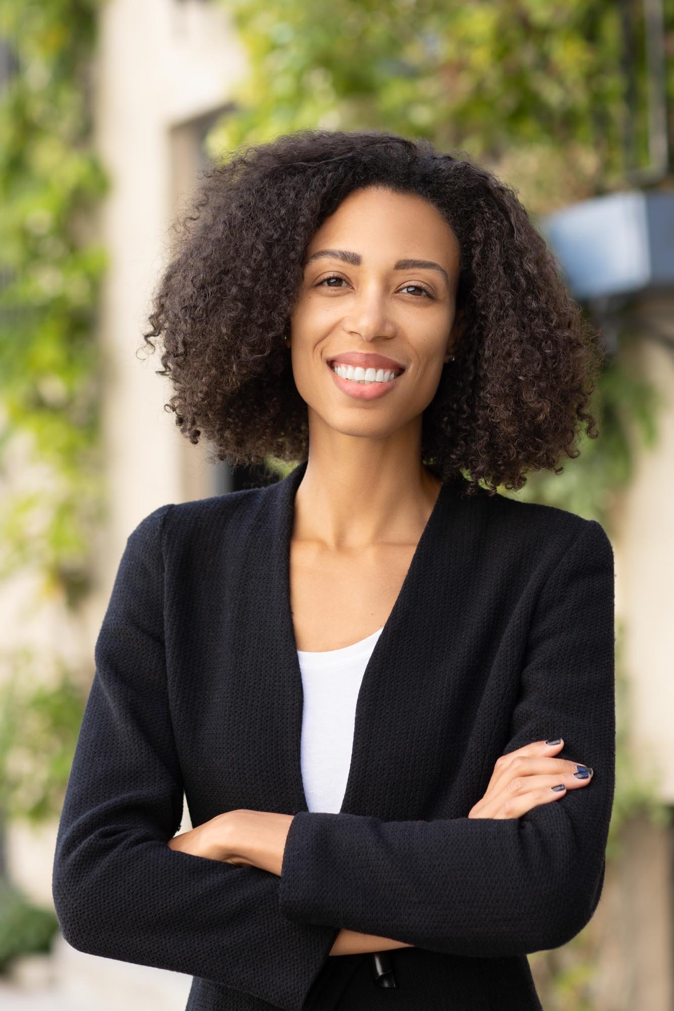 A woman with curly hair smiling outdoors with her arms crossed, wearing a black blazer and white shirt.