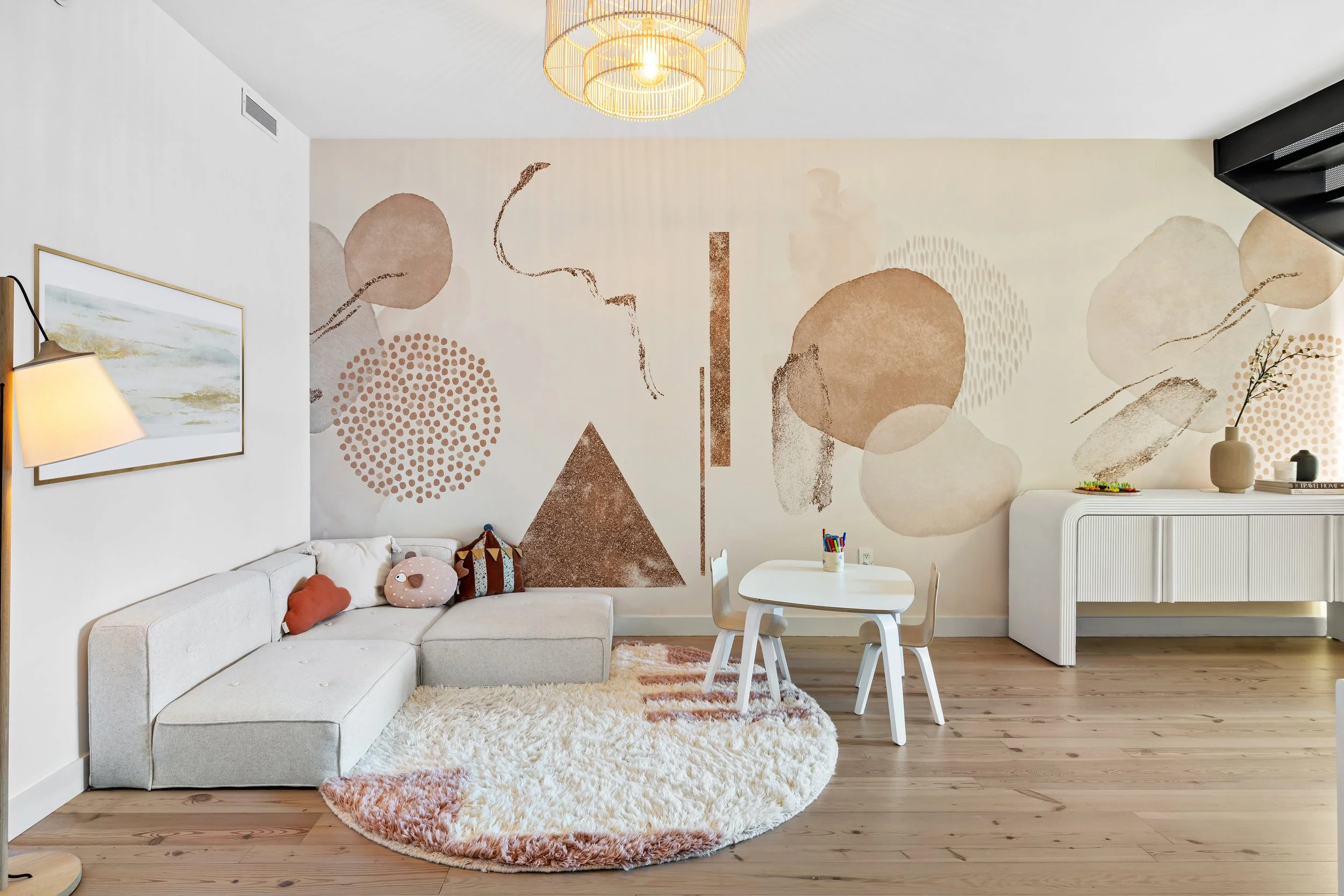 Contemporary living room with abstract wall art, a white sectional sofa with plush pillows, a circular shag rug, a small white children's table with chairs, and a white sideboard with decorative vases and books.