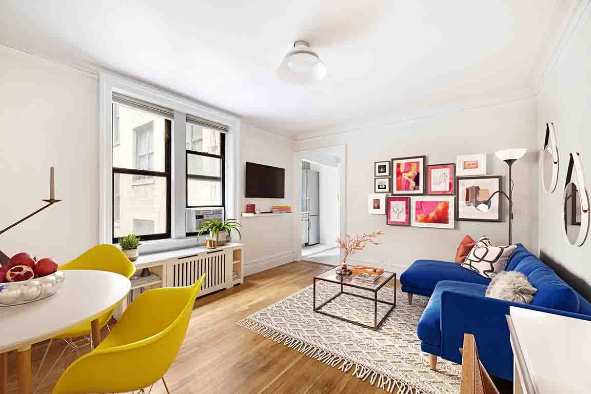 Bright living room with white walls, large windows, blue sofa, patterned rug, modern picture frames on the wall, and a dining table with yellow chairs.