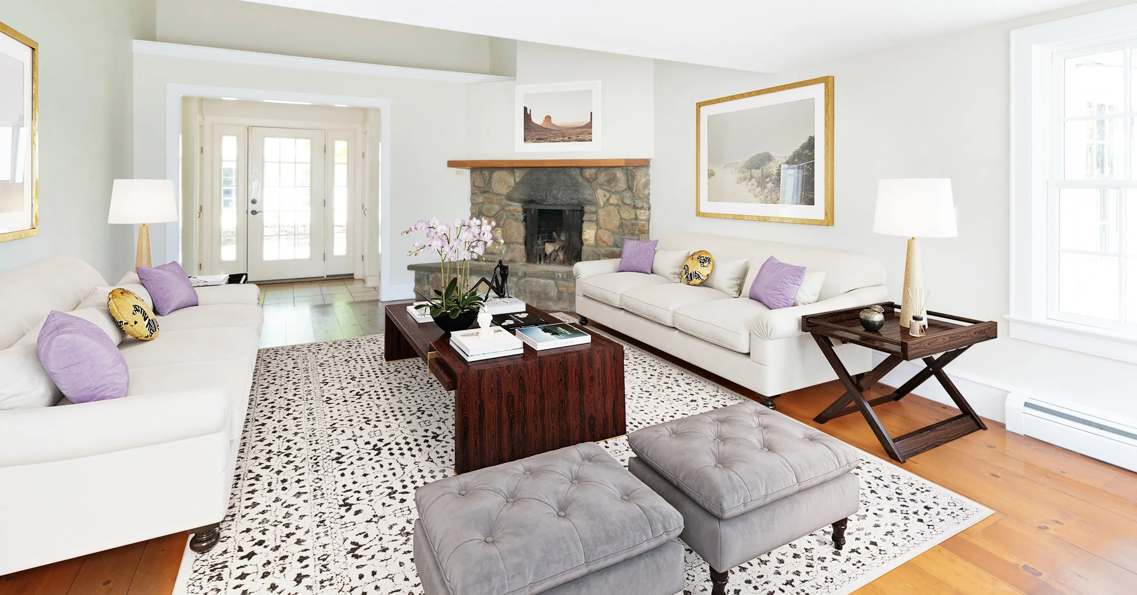 Bright living room with white sofas, purple and patterned cushions, a stone fireplace, wooden coffee table, two gray ottomans, decorative lamps, framed art, and large windows.
