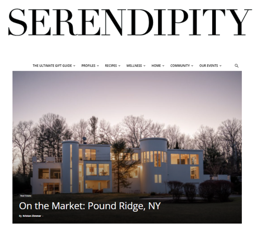 A modern white house with multiple levels and large windows, illuminated from within, set against a cloudy evening sky and surrounded by trees.