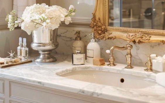 Bathroom sink area with white marble countertop, gold faucet, and decorative mirror. Items include a large silver vase with white flowers, soap dispenser, lotion bottles, a small box, and a candle.