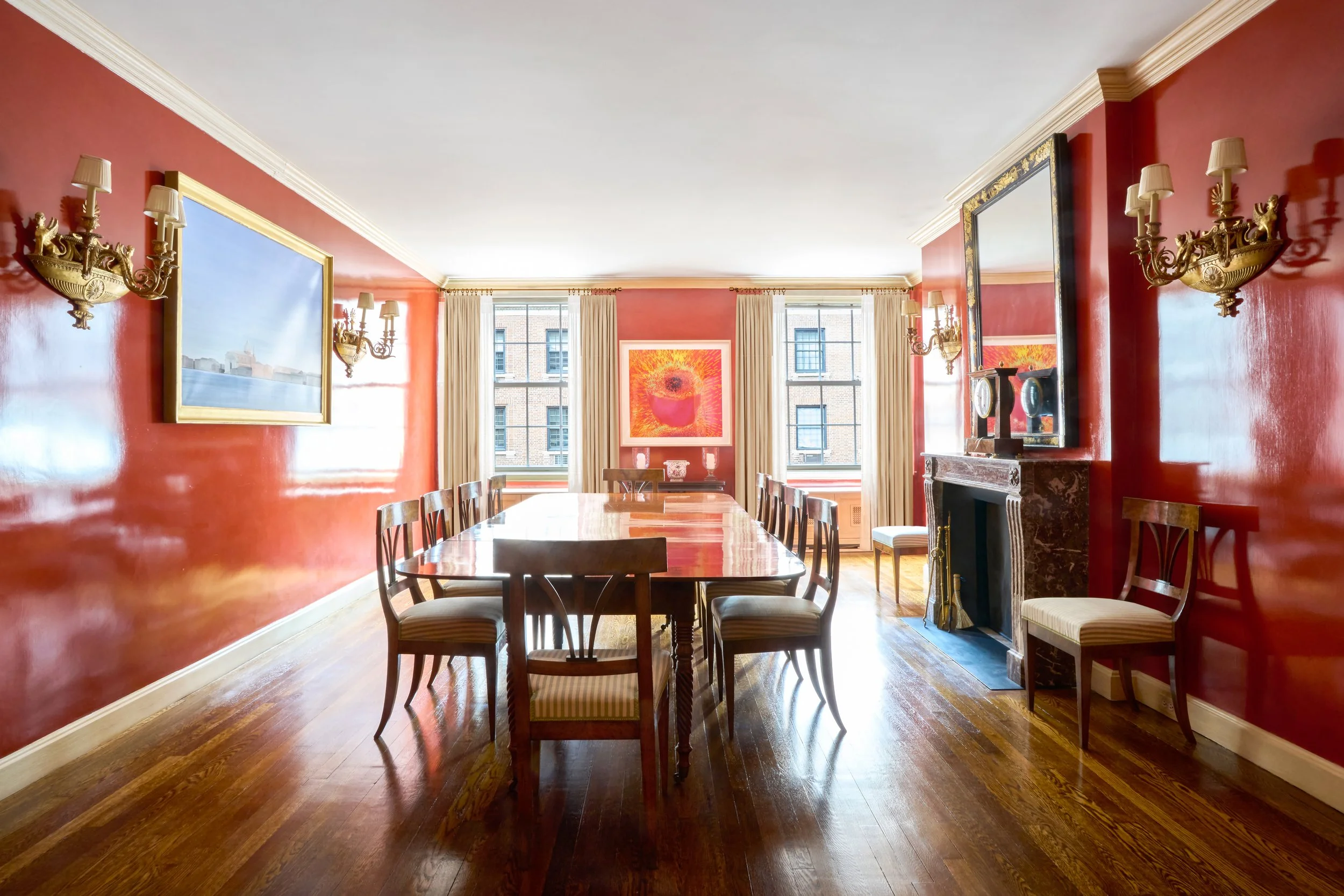 A spacious dining room with red walls, wooden floor, and a large rectangular wooden dining table with eight chairs. The room has two large windows with beige curtains, framed by artwork and decorative wall sconces. There's a fireplace with a mirror a