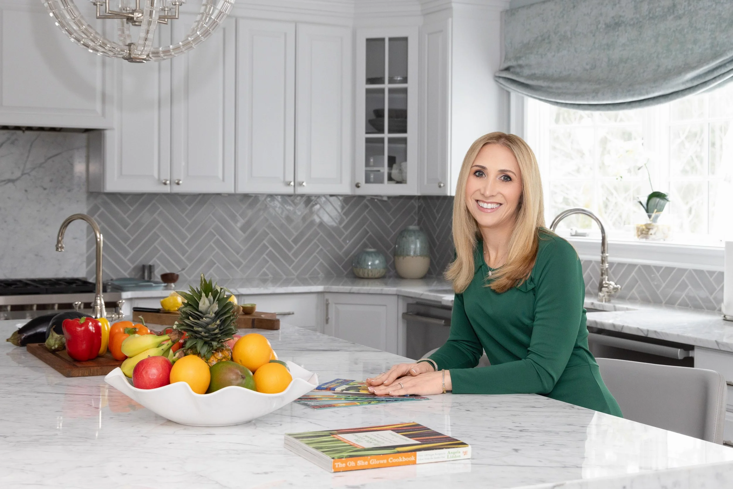 A woman in a green dress with blonde hair sitting at a kitchen island with a white bowl of assorted fruits including a pineapple, apples, oranges, bananas, and a mango, in a modern kitchen with white cabinets, gray backsplash, and large window.