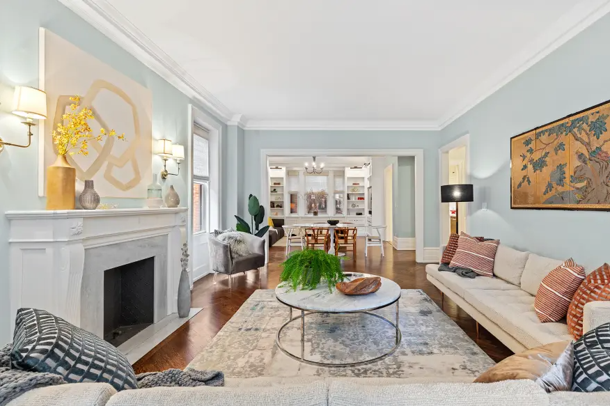 Living room with light blue walls, white fireplace, gray armchair, cream sofa with striped pillows, round coffee table with green plant, area rug, and built-in shelves in background.