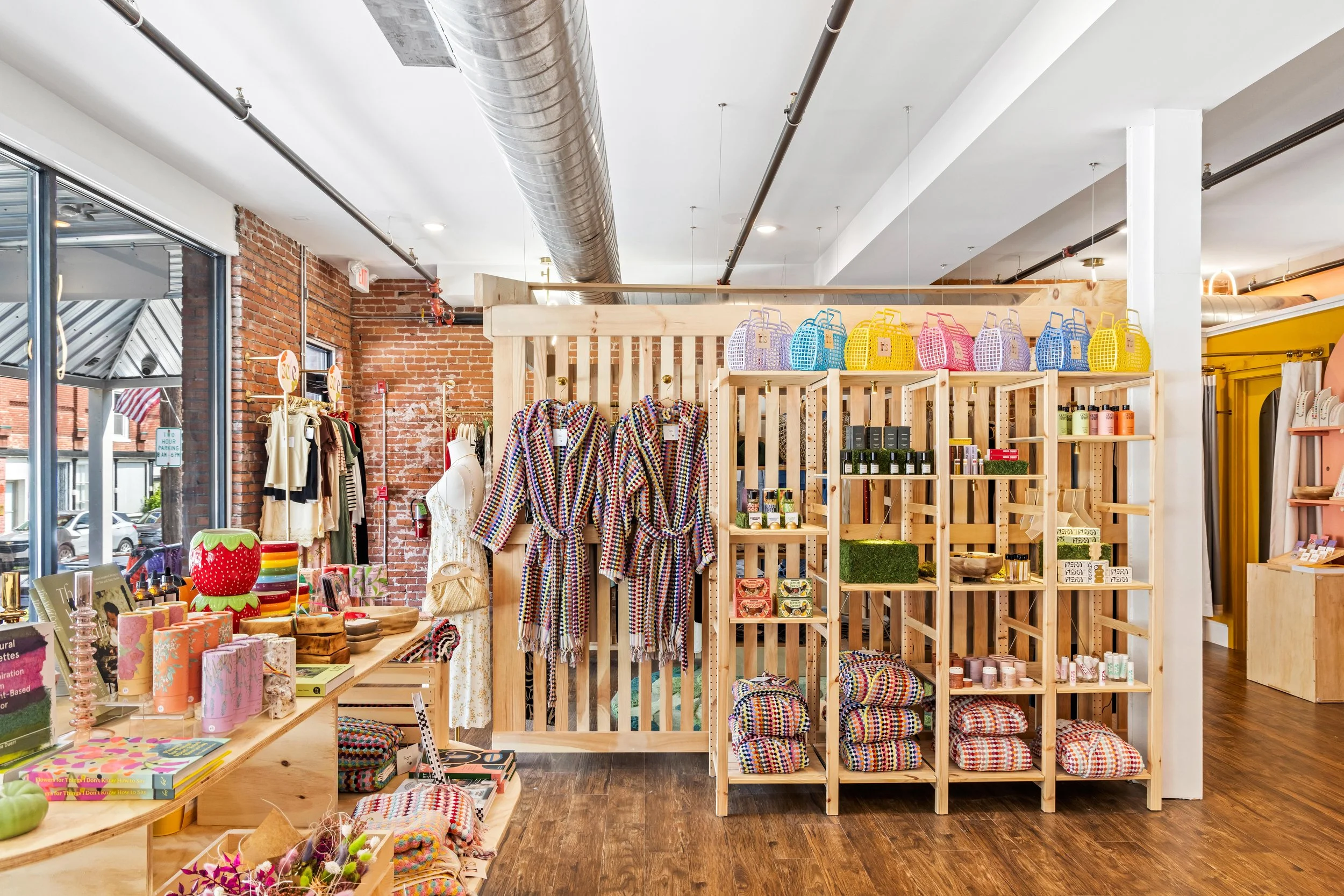 Interior of a boutique store with wooden shelving displaying colorful handbags, bath products, and folded blankets, with clothing racks and a table of various items, brick walls, and large windows.
