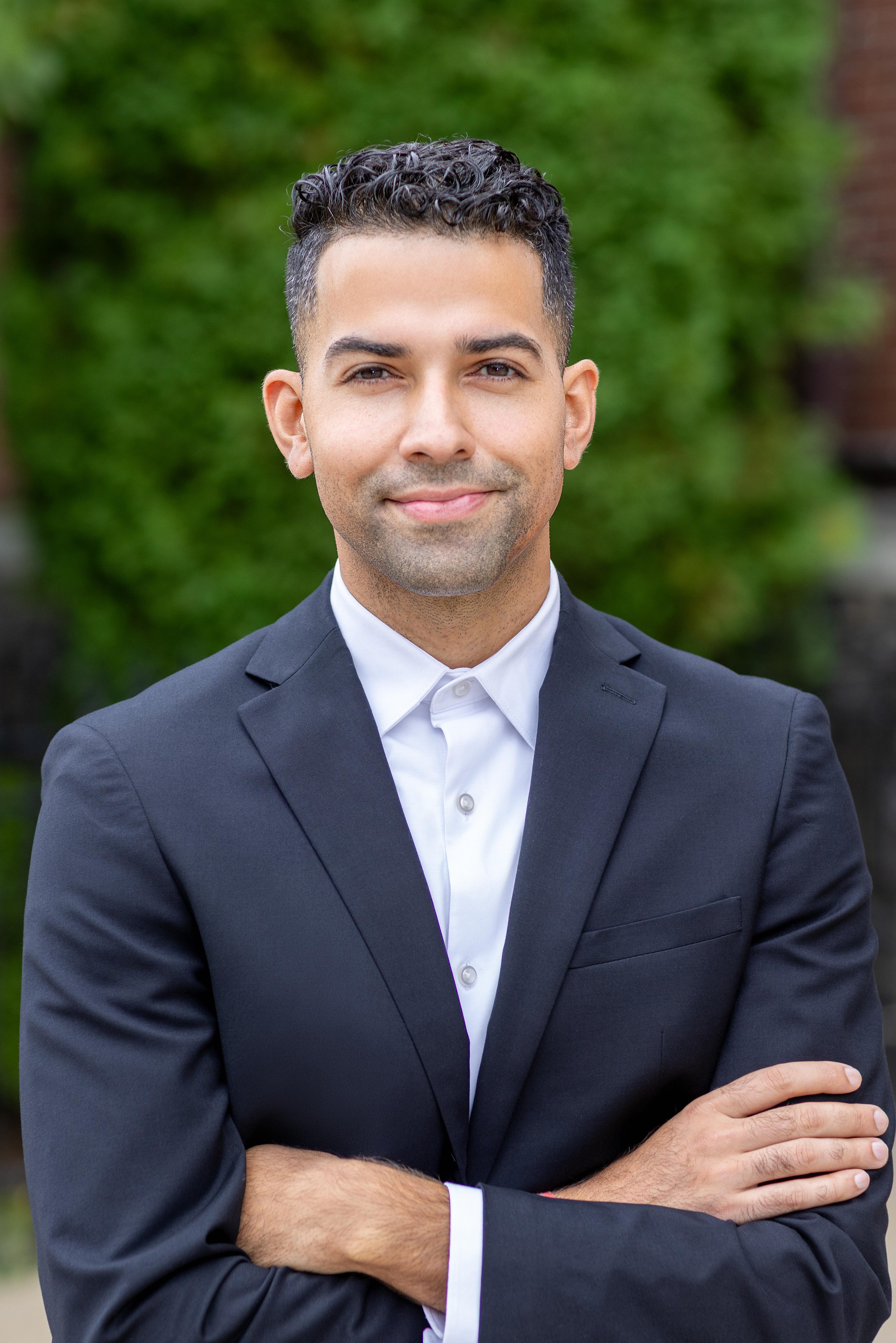 A headshot photo of man in a dark suit and white shirt standing outdoors with his arms crossed, smiling slightly, with greenery in the background.