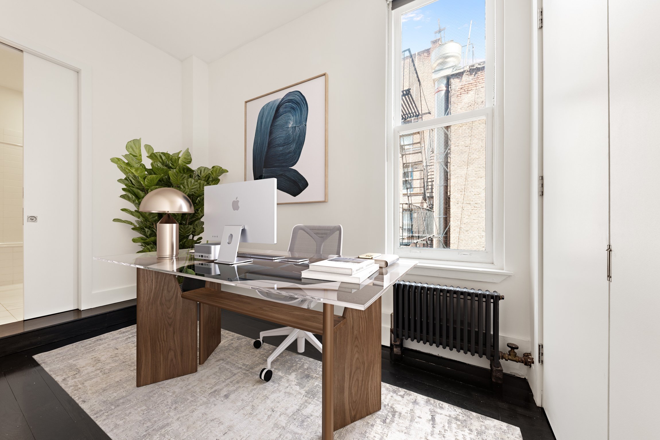 Modern office with a desk, iMac computer, lamp, books, and a gray ergonomic chair, next to a large window, with a black radiator below.