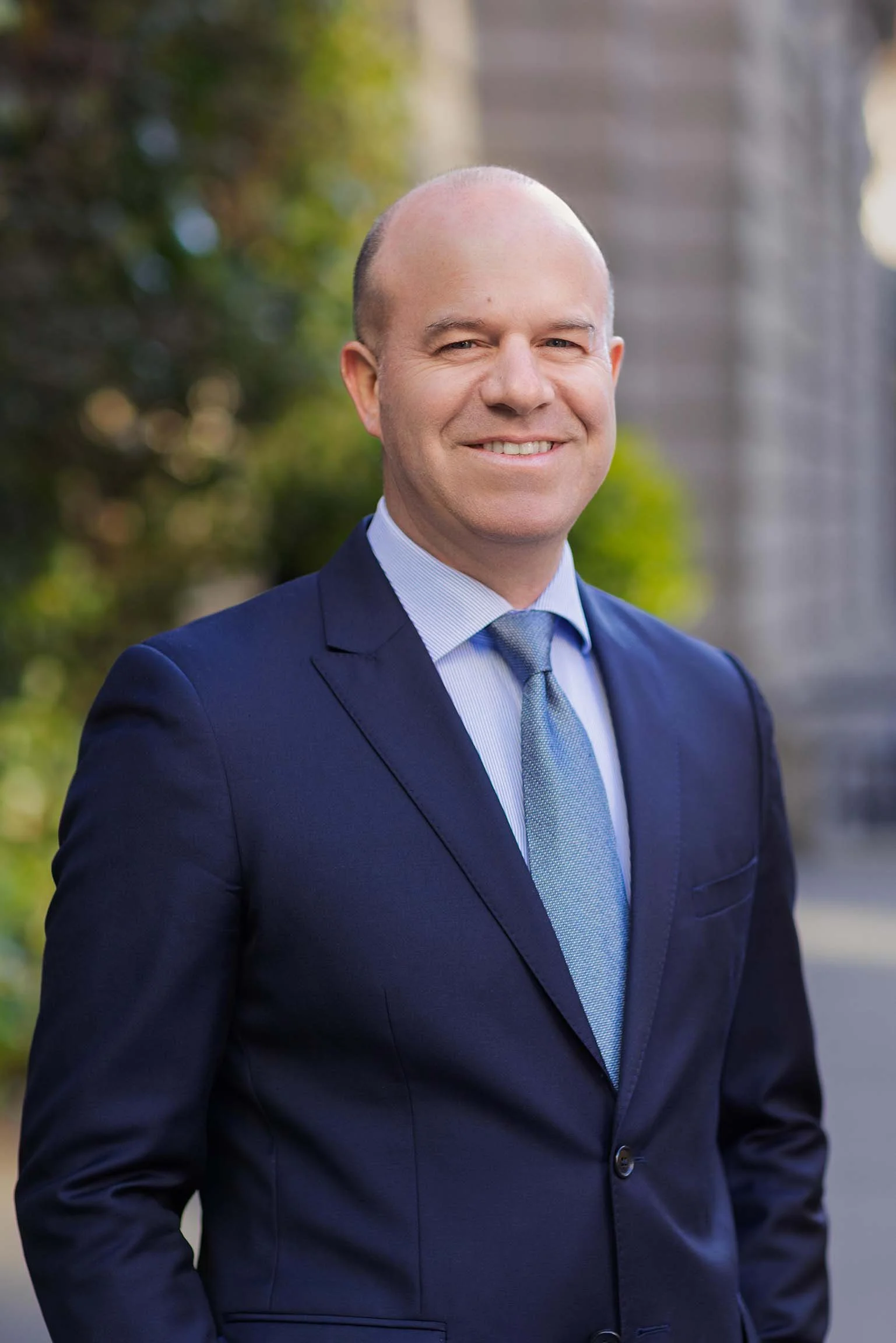 A man in a blue suit and tie smiling outdoors, with blurred greenery and a building in the background.