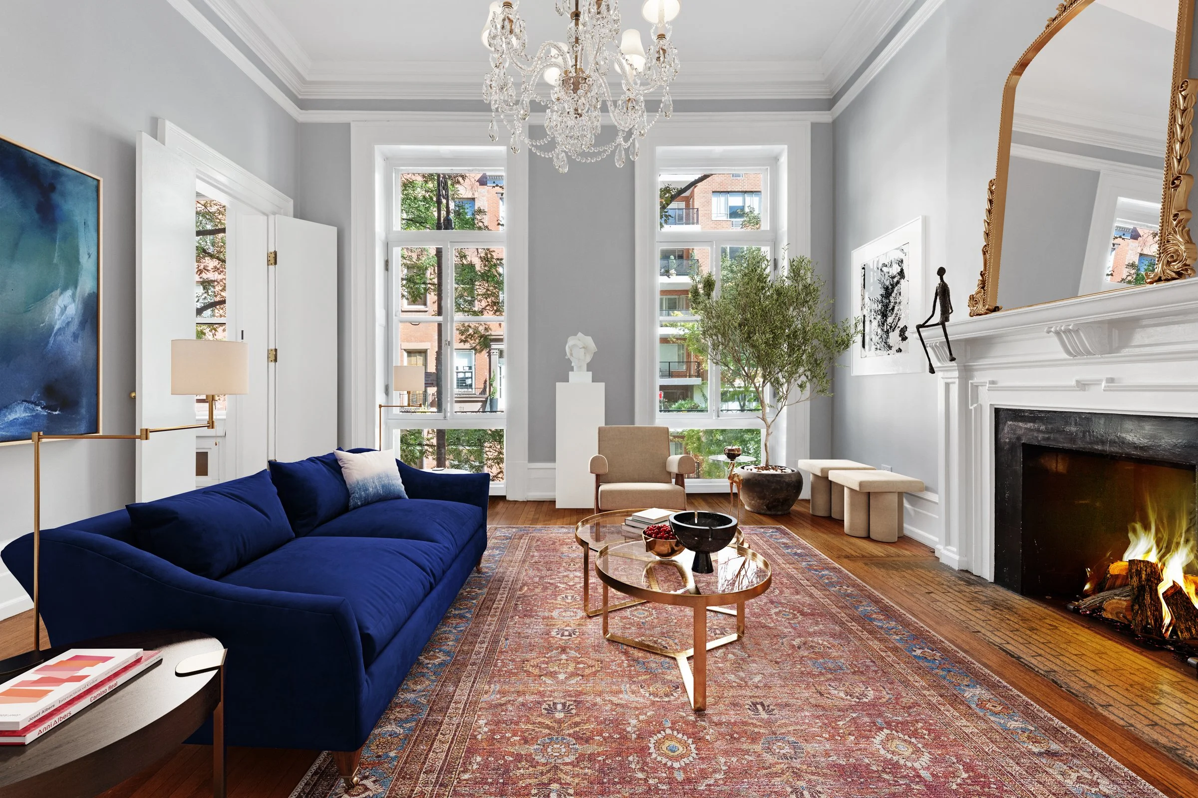 Elegant living room with large windows, a fireplace with a fire, a blue velvet sofa, a beige armchair, a vintage-style rug, a chandelier, and decorative art and sculptures.
