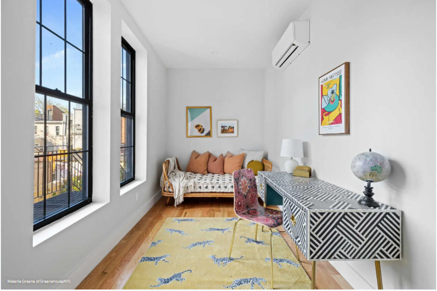 Bright room with three large windows, a white wall, and hardwood floors. There is a patterned couch with multiple cushions, a colorful chair, a black and white patterned sideboard with a globe and lamp, and framed artwork on the walls.