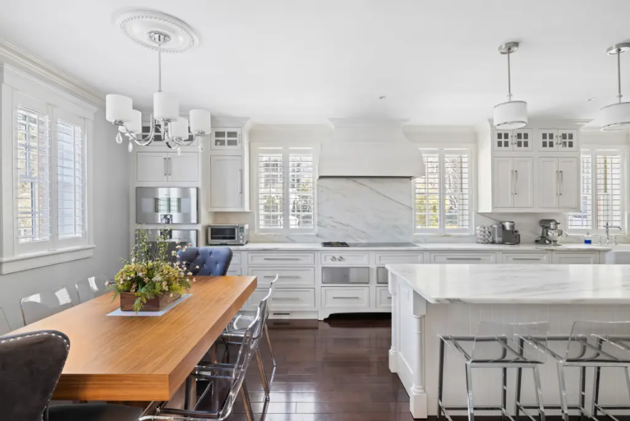 Bright white kitchen with large marble island, wooden dining table with flowers, and multiple windows with shutters.