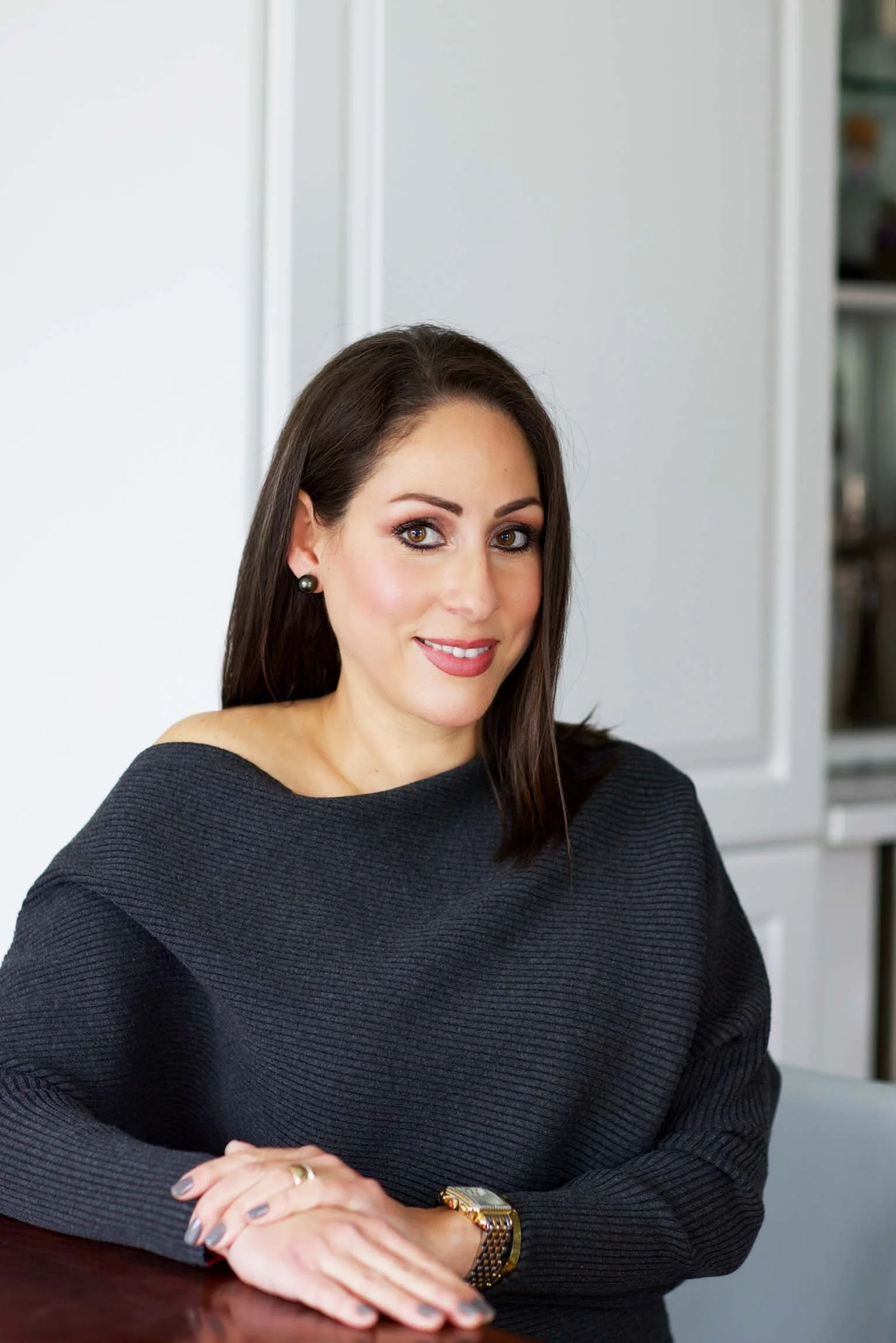 A woman with long dark hair, wearing dark earrings and a dark sweater, sitting at a table in a modern kitchen or office, smiling at the camera.