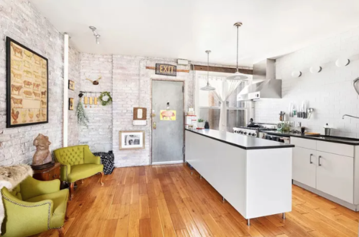 Modern kitchen with white brick walls, wooden flooring, and a white island. Decor includes framed artwork, a green vintage chair, and hanging pendant lights.