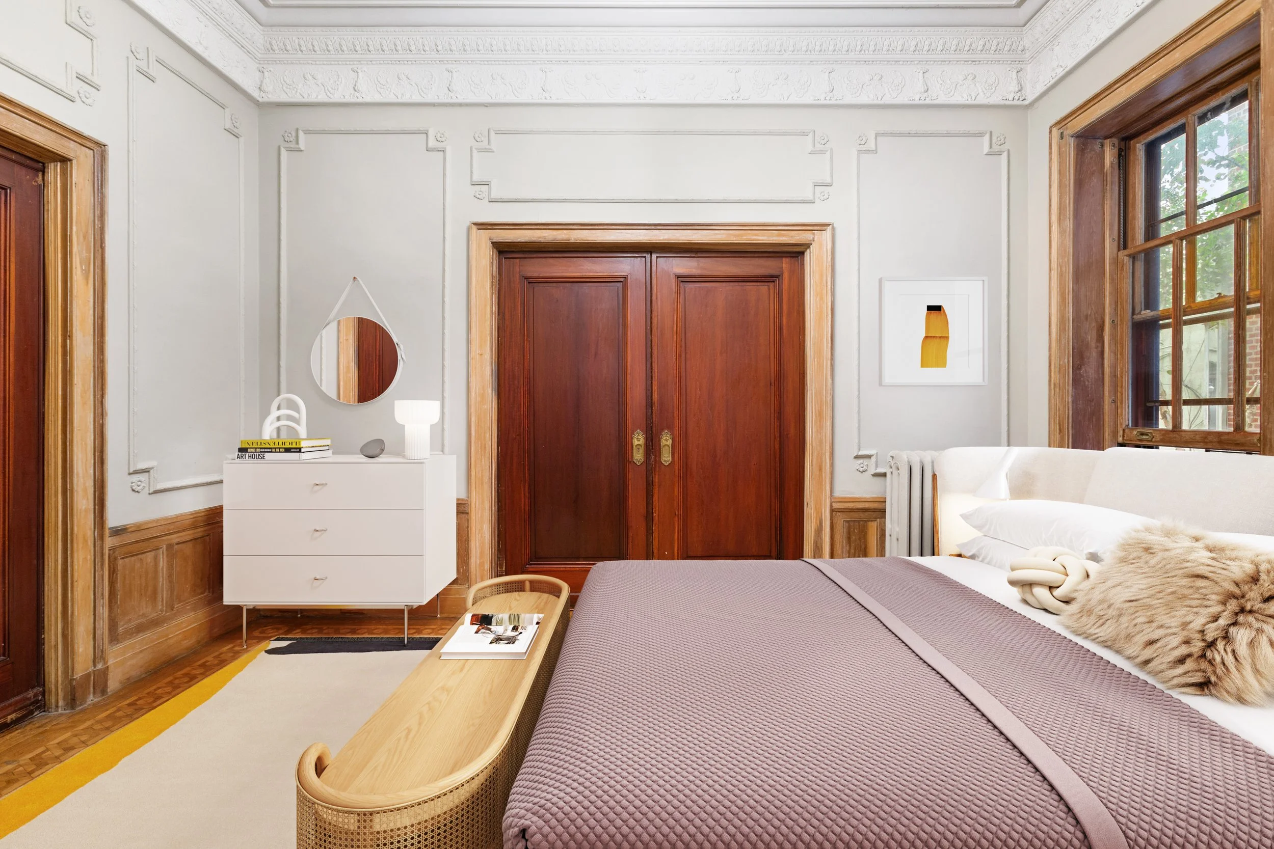 A bedroom with white walls, wooden trim, a bed with a light purple quilt, and a window with wooden frames. A white dresser with decorative items is to the left of the wooden closet doors. There is a wall art piece on the right wall and a mirror hangi