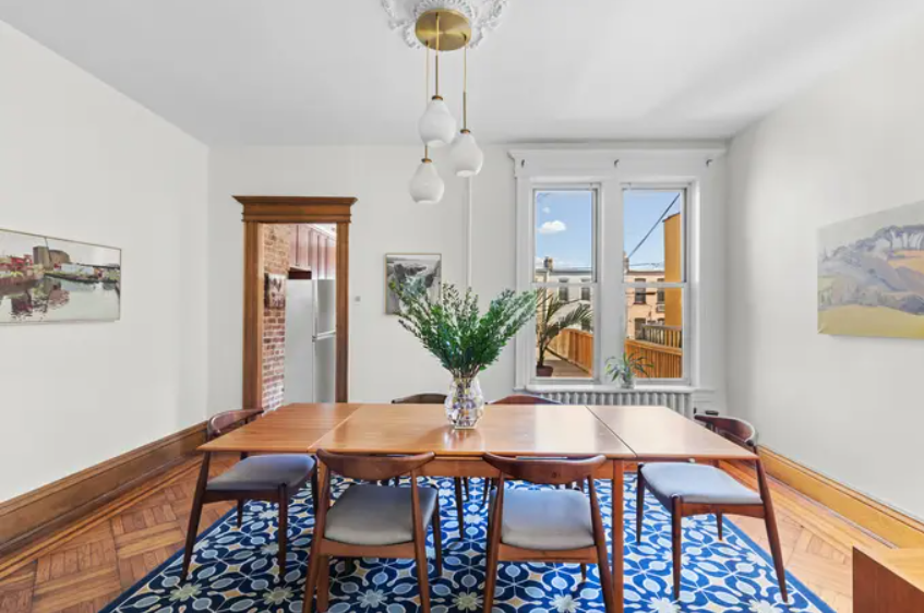 Bright dining room with large window, wooden table, six chairs, pendant light fixture, patterned rug, and artwork on white walls.