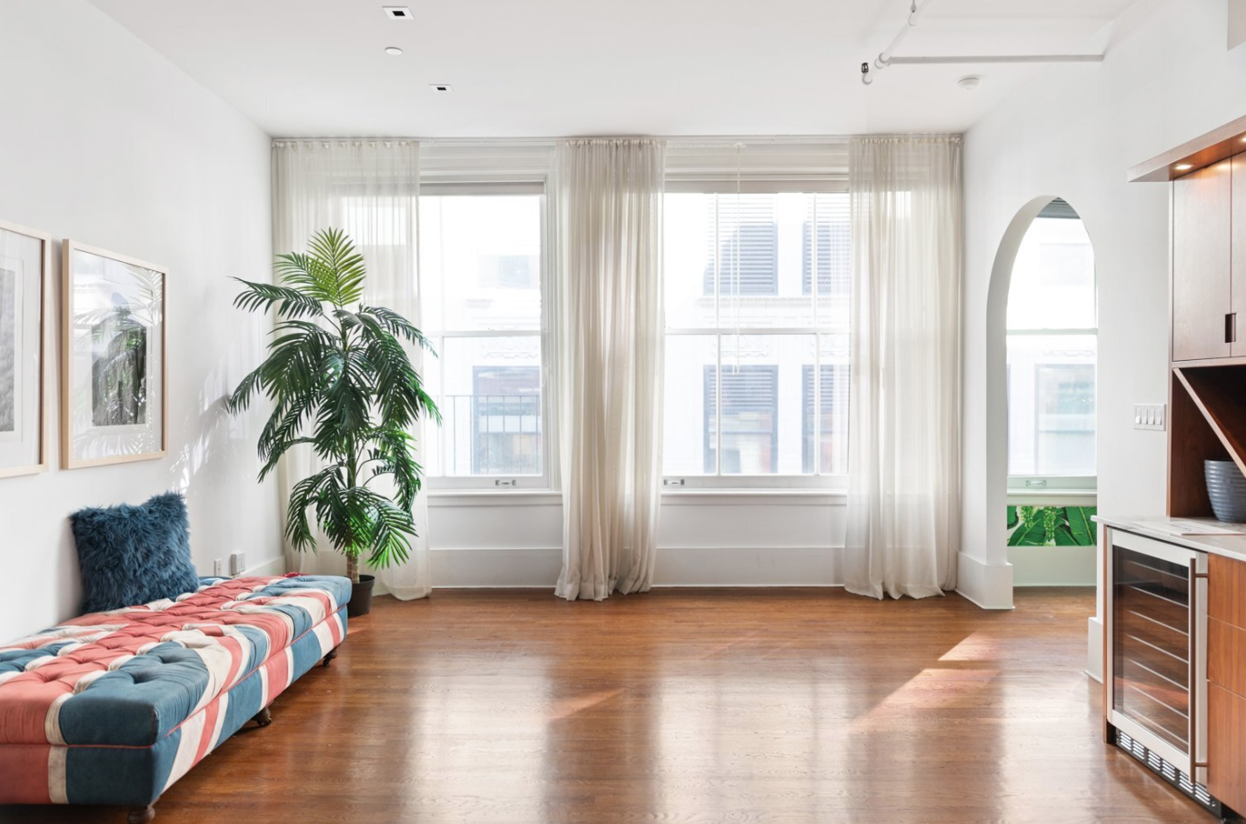 Bright living room with large windows, sheer white curtains, a tall green plant, a striped bench with a blue pillow, framed artwork on the wall, and wooden flooring.