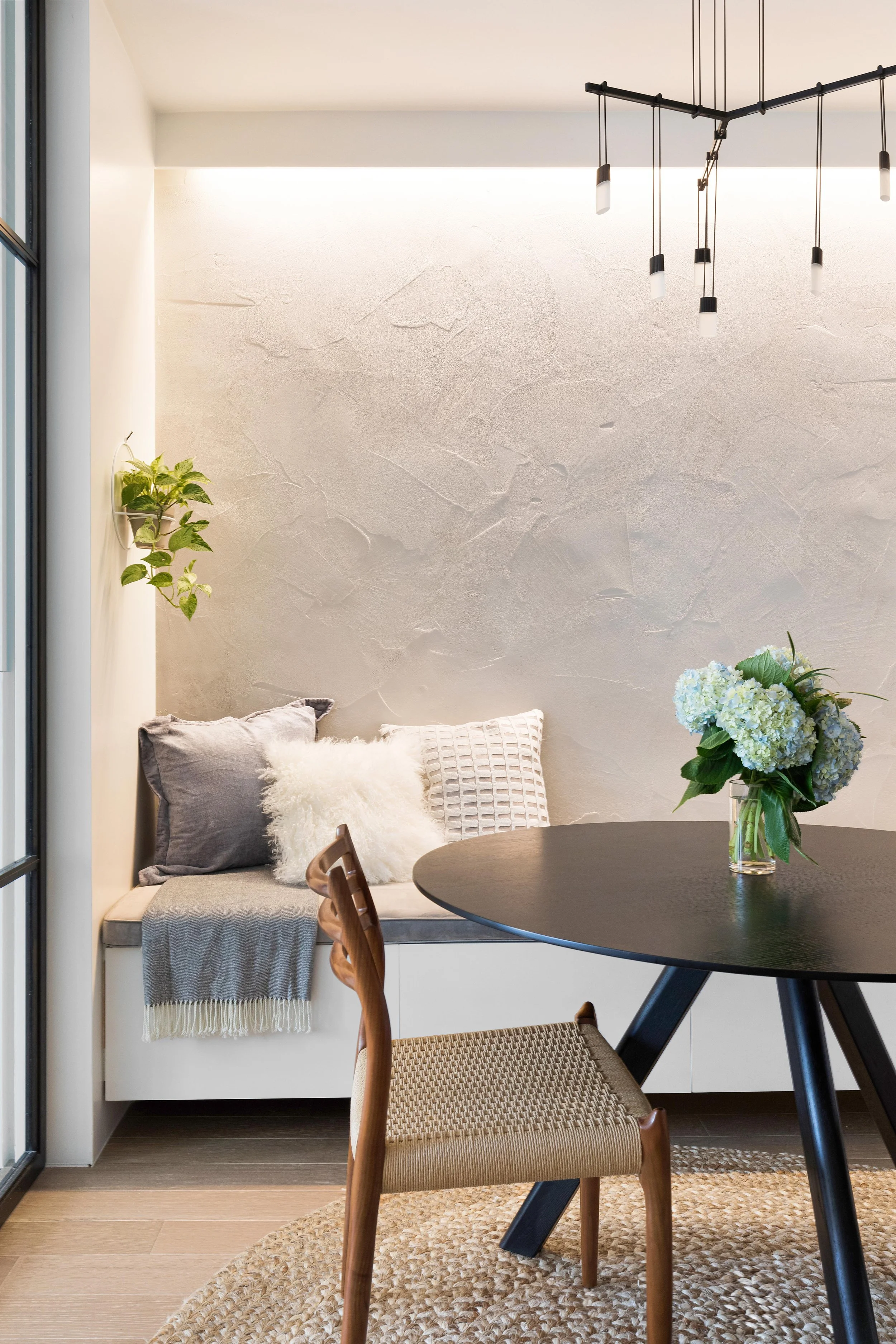 Cozy dining nook with a round black table, wooden chair, and a window seat with pillows and a blanket. A vase with white hydrangeas sits on the table, and a hanging light fixture is above. A small green plant is on the wall.