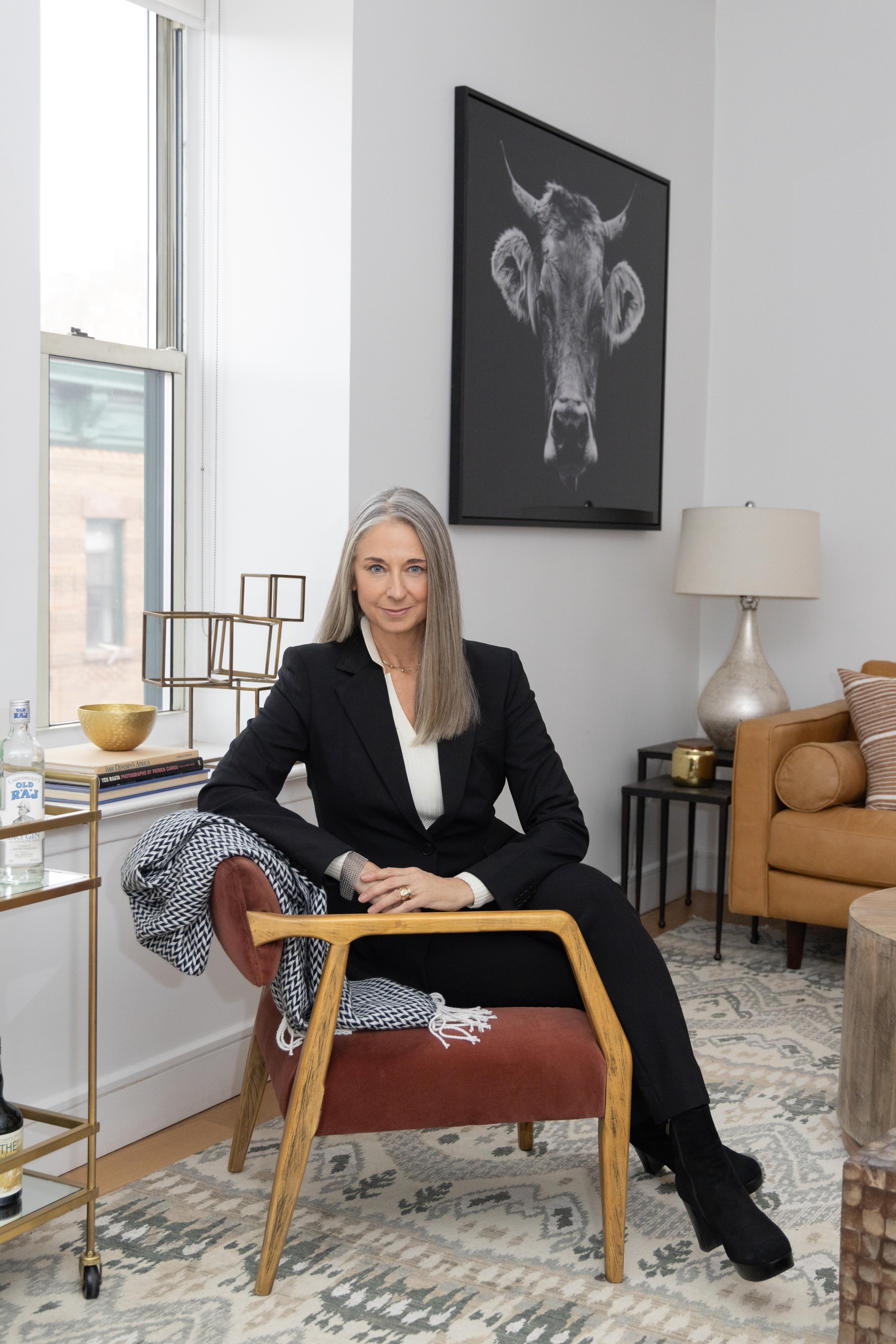 A woman with long gray hair sitting on a red chair in a modern living room, dressed in a black suit. The room has a large window, a white wall with a black and white cow portrait, a beige sofa with brown cushions, a black side table with a lamp, and 