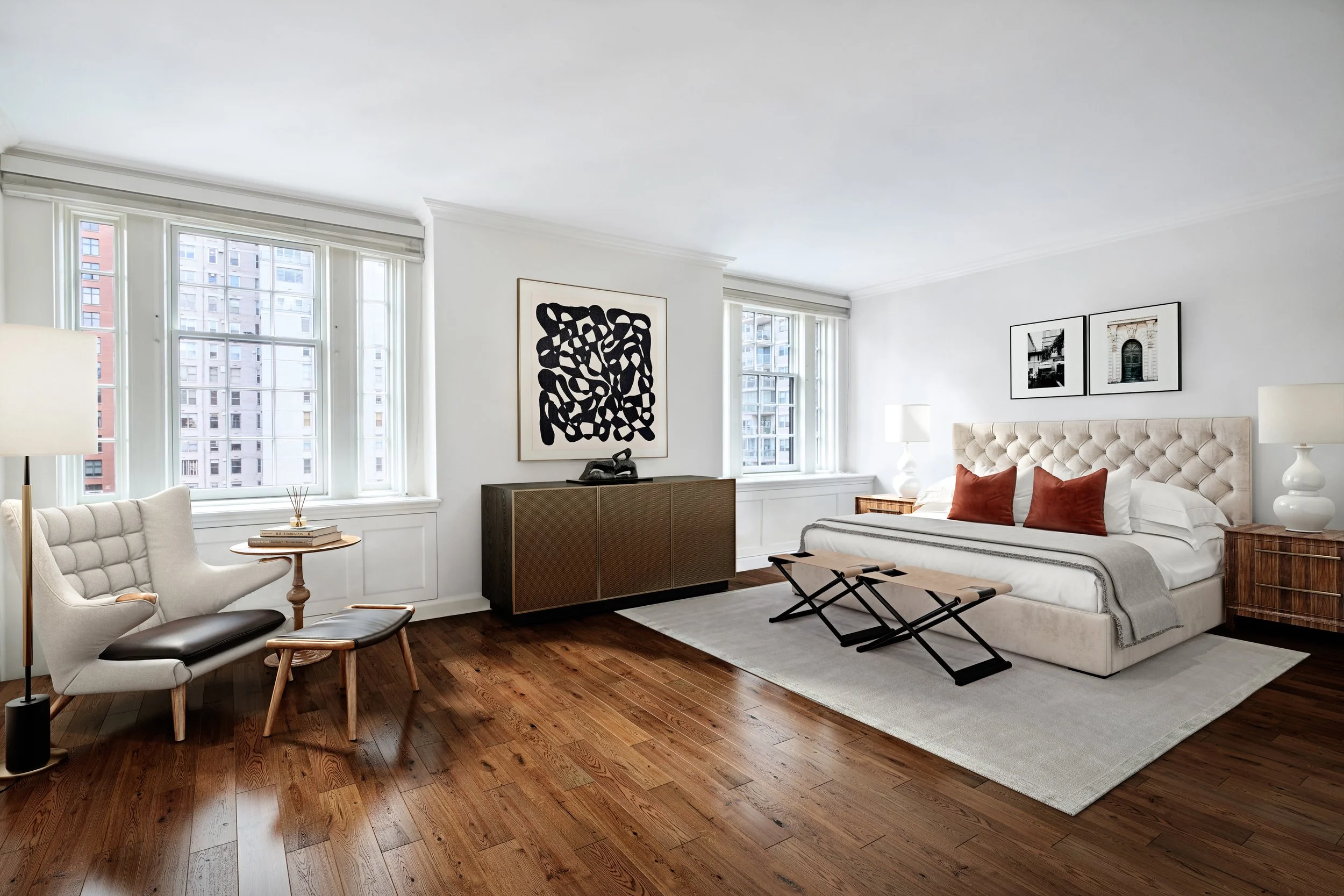 Bright modern bedroom with a large bed, tufted headboard, two nightstands with lamps, three framed pictures above the bed, a white armchair with a matching footstool, a side table with books, wooden flooring, two additional black stools at the foot o