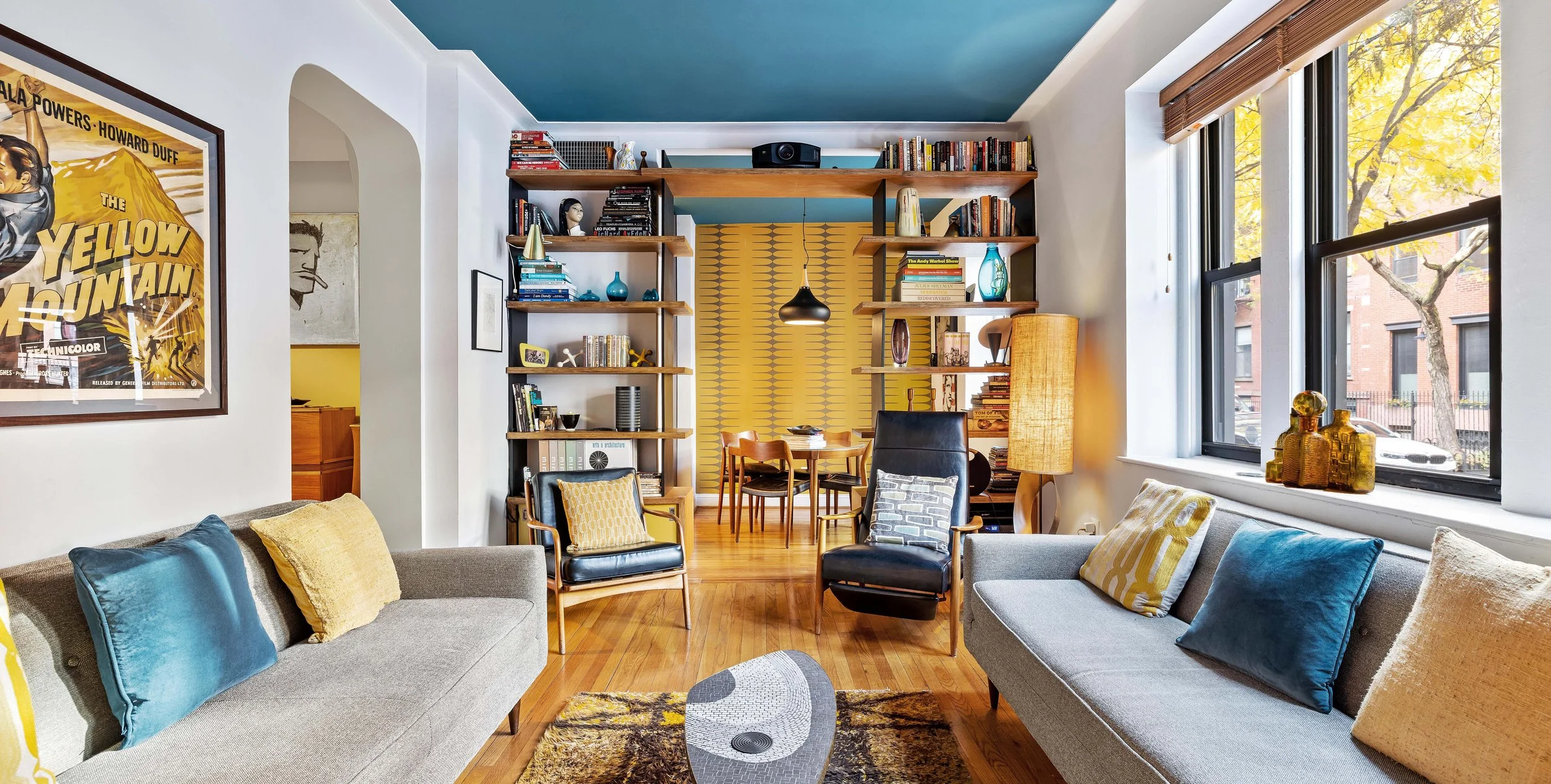 Living room with two gray sofas with colorful pillows, two black armchairs with pillows, a small oval coffee table, large windows with view of trees and street, open bookshelf with books and decorative items, yellow accent wall, and dining area with 