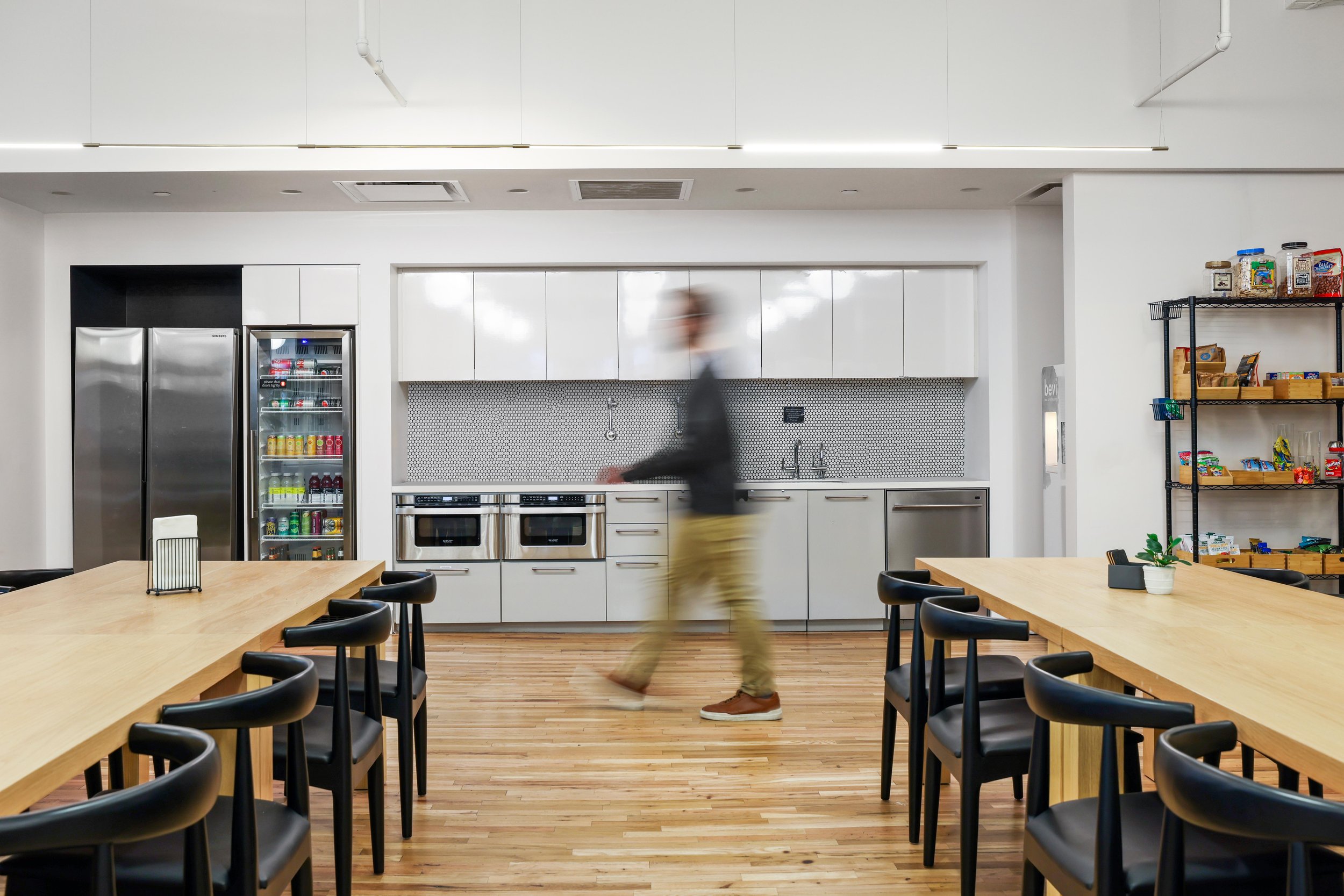 A person walking in a modern office kitchen, with black chairs around wooden tables, a stainless steel refrigerator, a beverage cooler, white cabinets, and a black metal shelving unit with snacks and supplies.