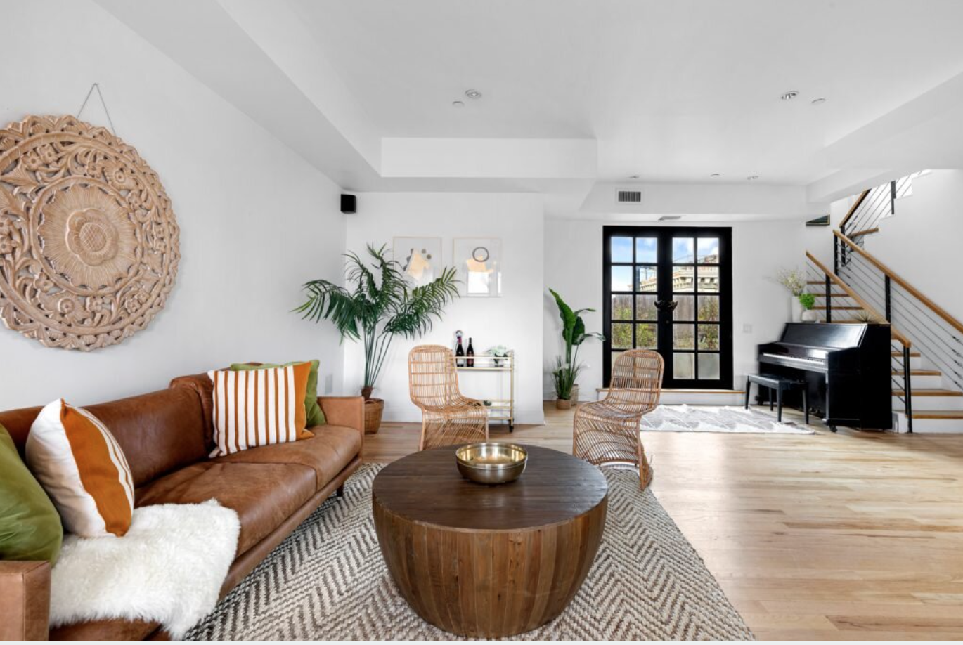 Living room with a leather sofa, striped pillows, wicker chairs, a round wooden coffee table, potted plants, black double doors, a black piano, and staircase with wooden handrail.
