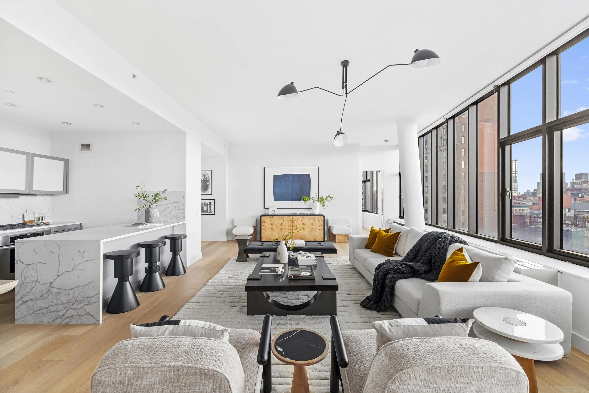 Modern apartment living room with white walls, large windows, black and white furniture, yellow accent pillows, black coffee table, and wall art.