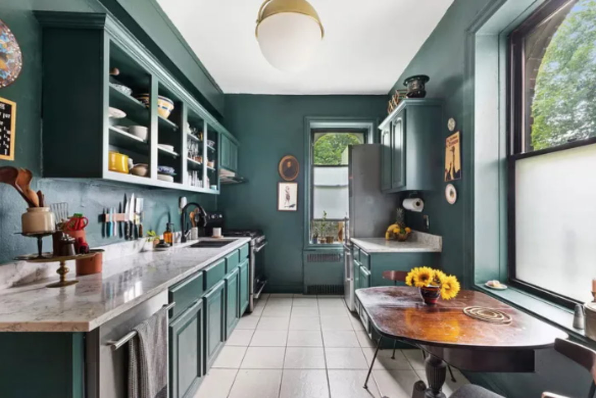 A kitchen with teal cabinets, a marble countertop, and a round wooden dining table with a vase of sunflowers. There are open shelves displaying dishes, a window with green trees outside, and various kitchen appliances and decor.