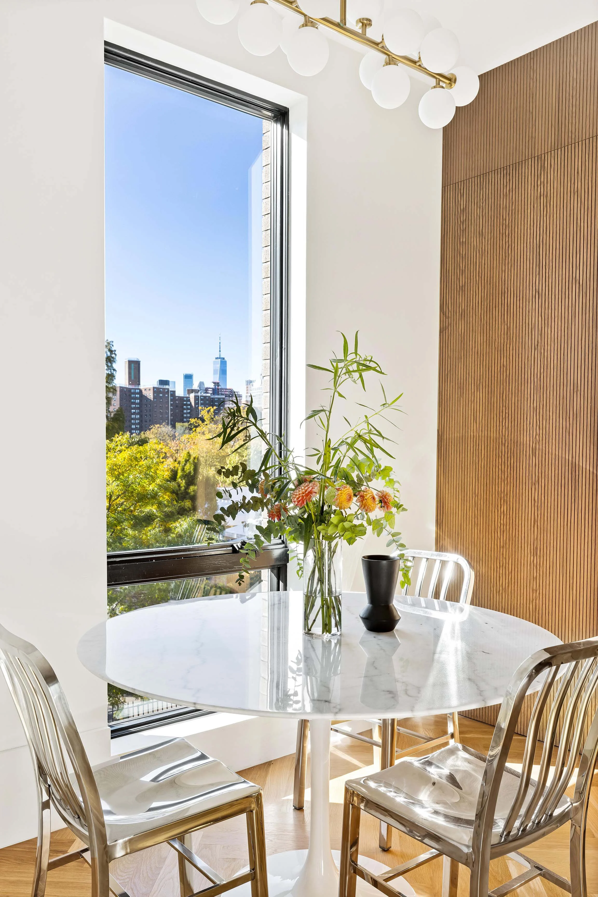 Modern dining area with a white marble round table, four metallic chairs, a large window showing a city skyline and trees, a tall glass vase with flowers, a black vase, and a wood-paneled wall.