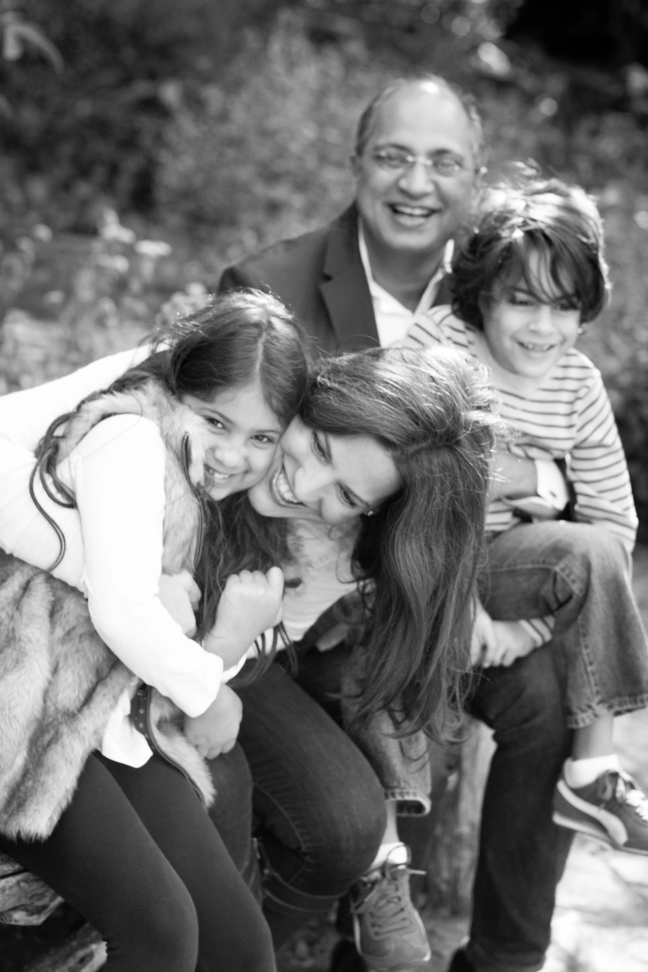 A group of four people, two adults and two children, smiling and enjoying each other’s company outdoors, sitting on a bench in a park.
