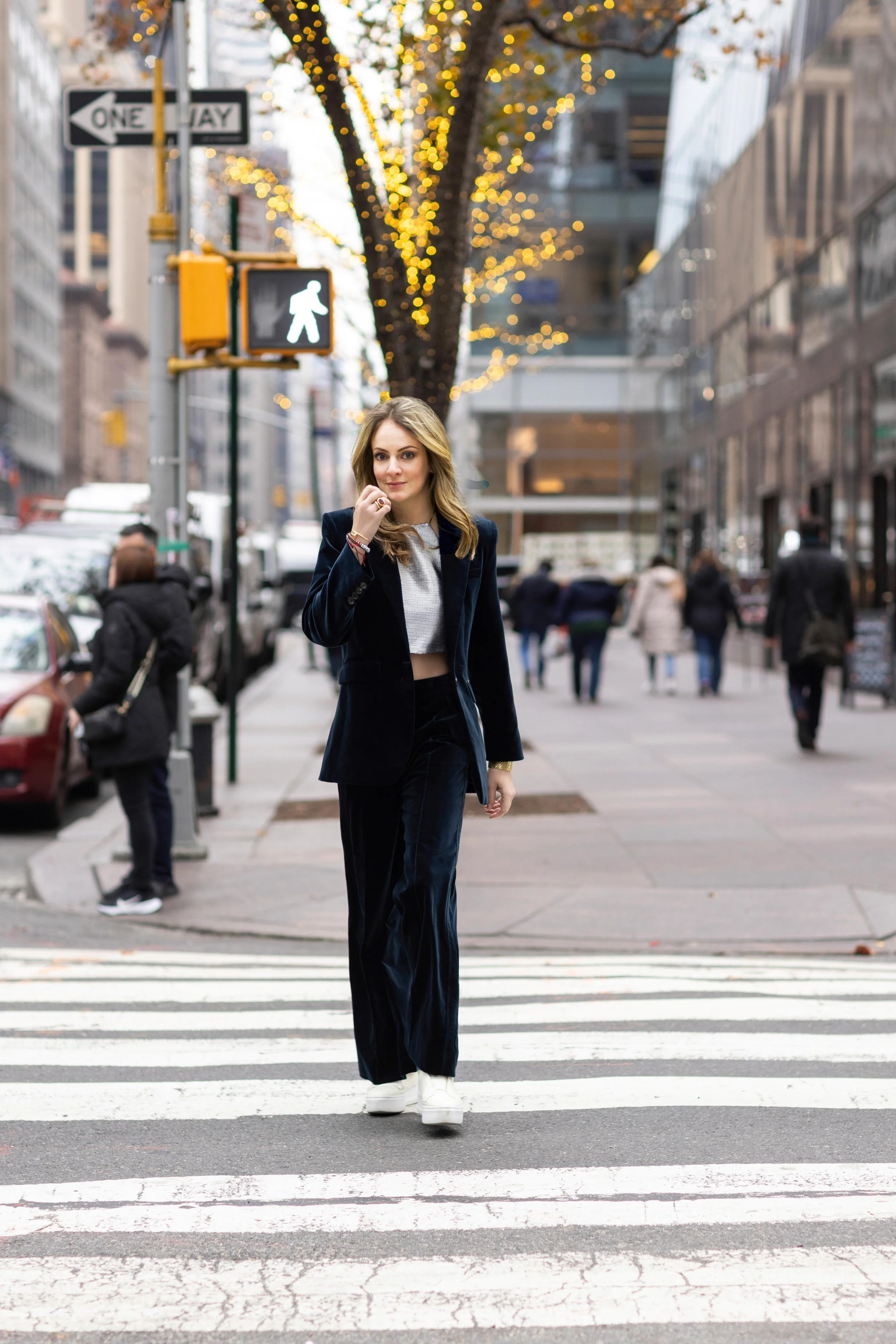 A woman walking on crosswalk in an urban city at dusk. She is dressed in a black velvet suit with white platform shoes. Pedestrians are visible in the background, and the street is decorated with string lights on a tree. A pedestrian crossing light s