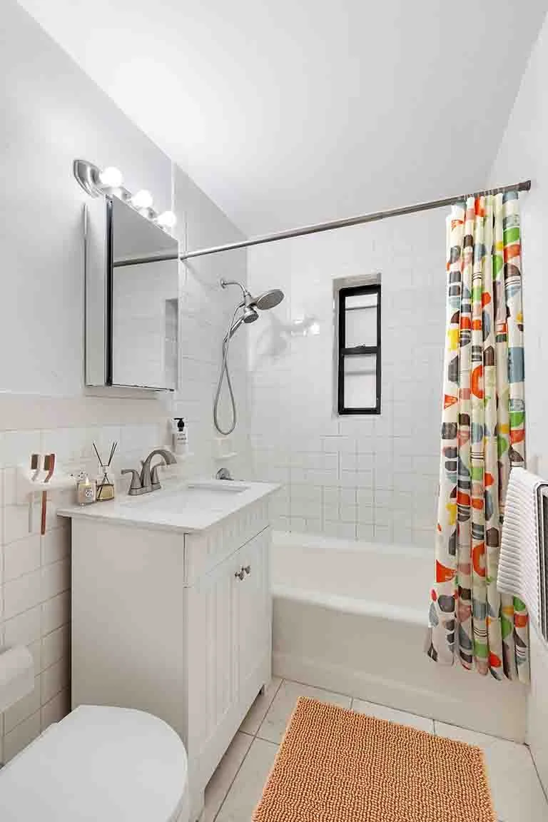 Small bathroom with a white vanity, a mirror with lights above, a bathtub with a showerhead, a colorful shower curtain, a small window, and a towel hanging on a rack.