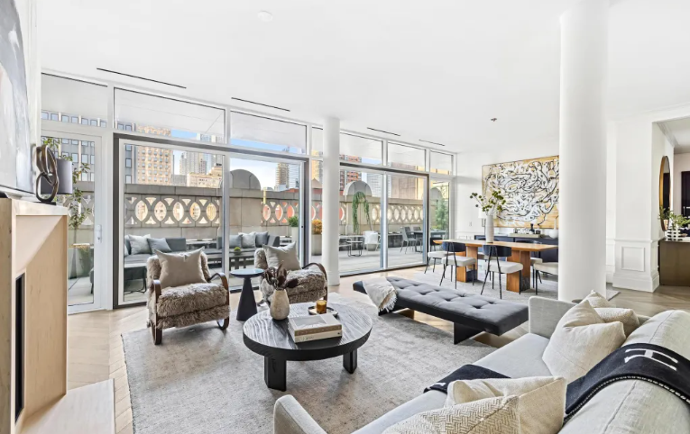 Modern living room with large windows, gray and beige furniture, coffee table, and balcony with outdoor seating, city view.