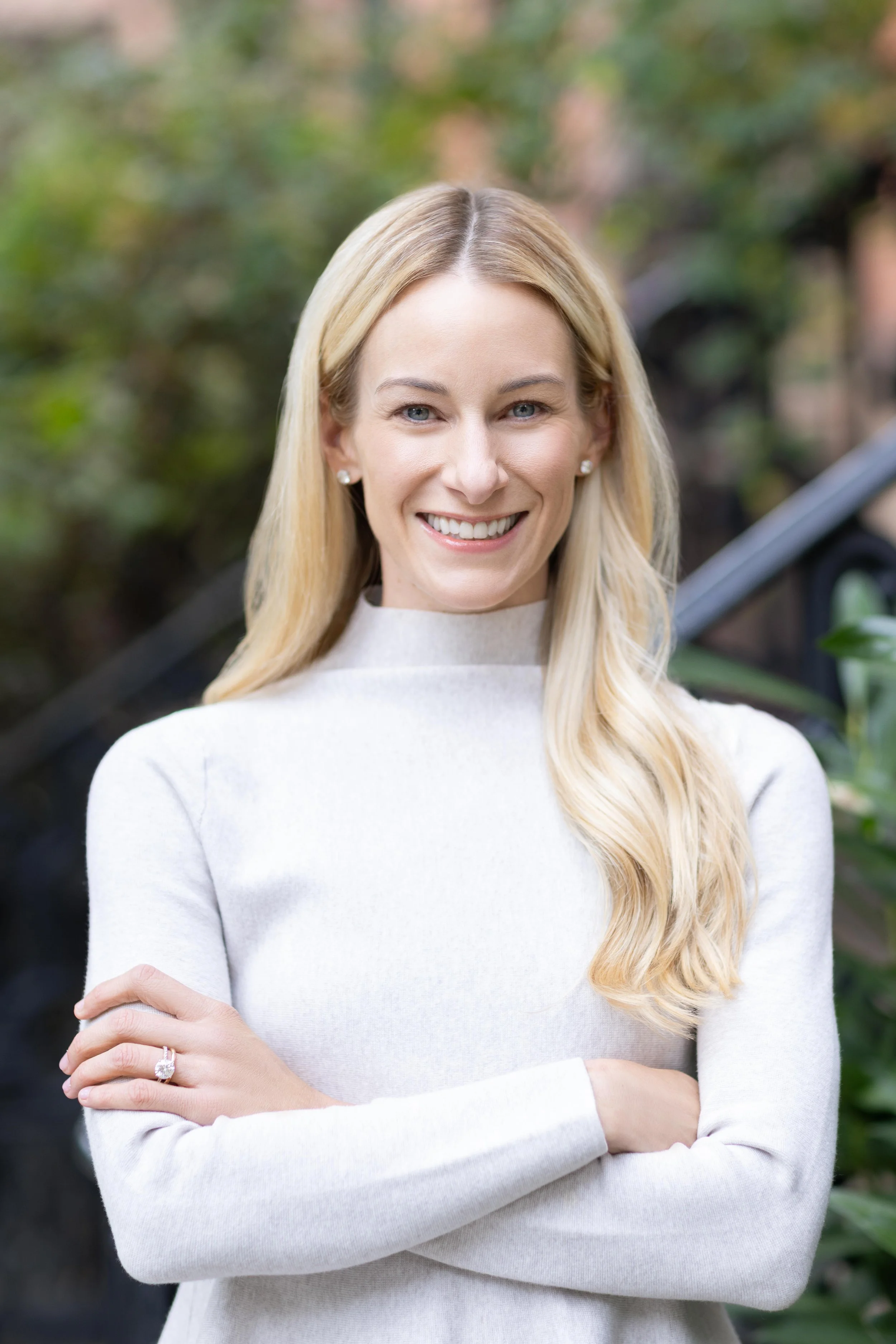 A smiling blonde woman with crossed arms standing outdoors in front of a blurred background of greenery.