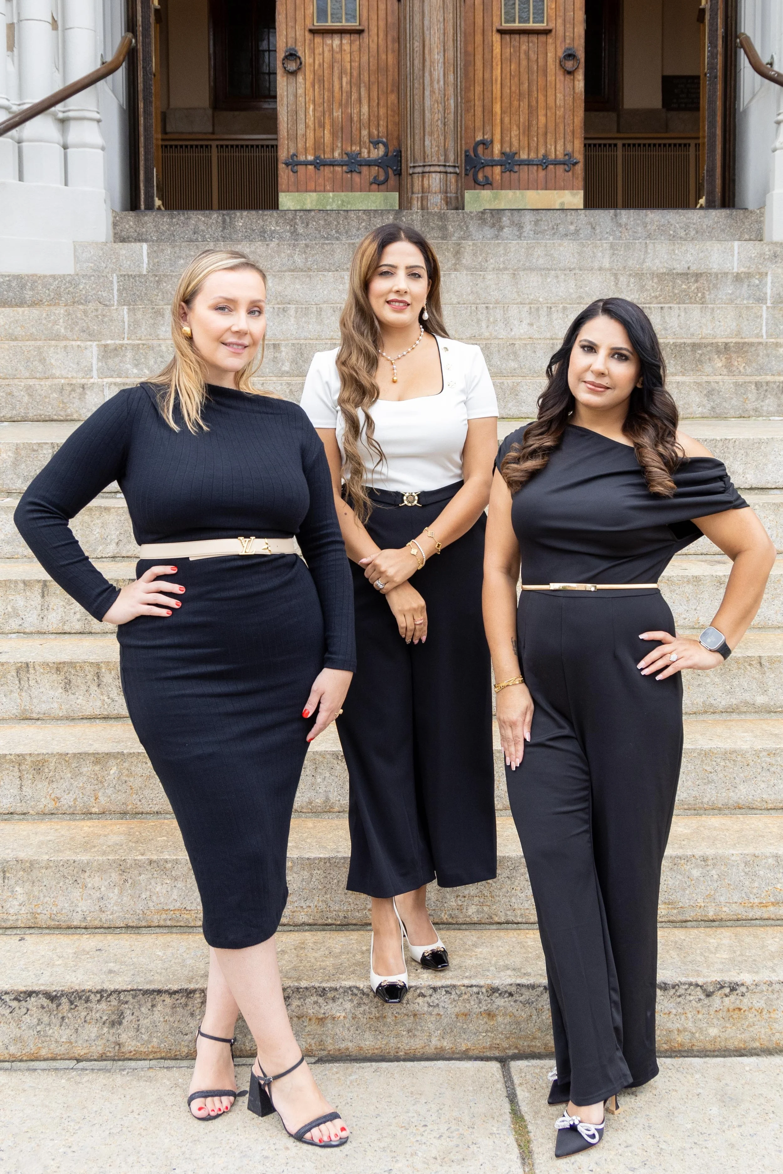 Three women in formal black and white outfits standing on outdoor steps in front of a wooden door.