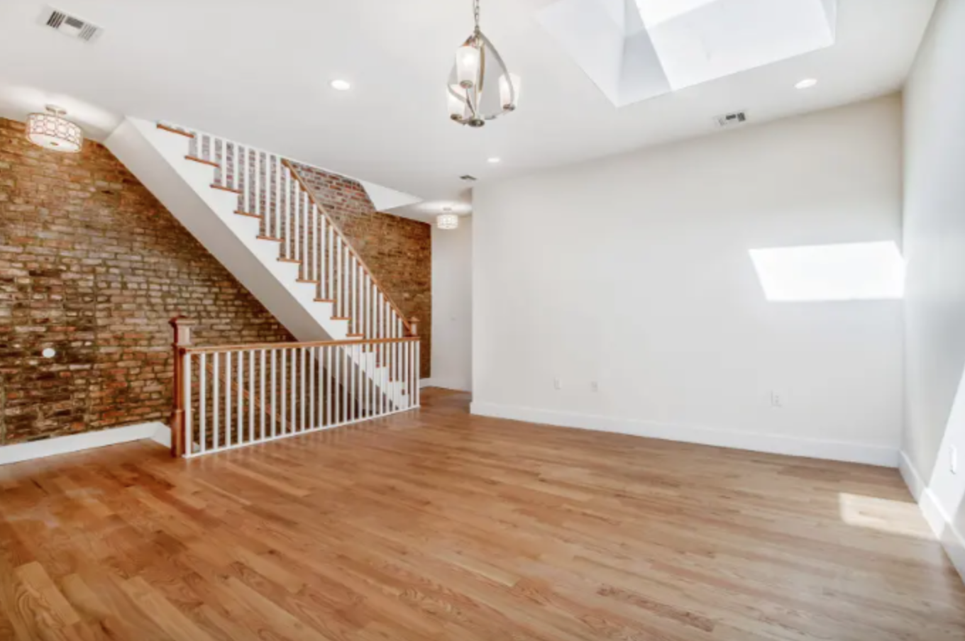 Empty living room with hardwood floors, white walls, a staircase with a brick wall background, skylights, and modern lighting fixtures.