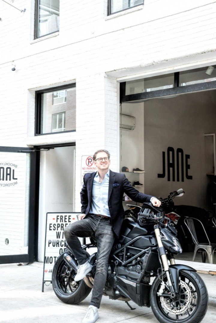 A man in a blazer and jeans sitting on a black motorcycle outside a white building with a sign indicating a coffee shop.
