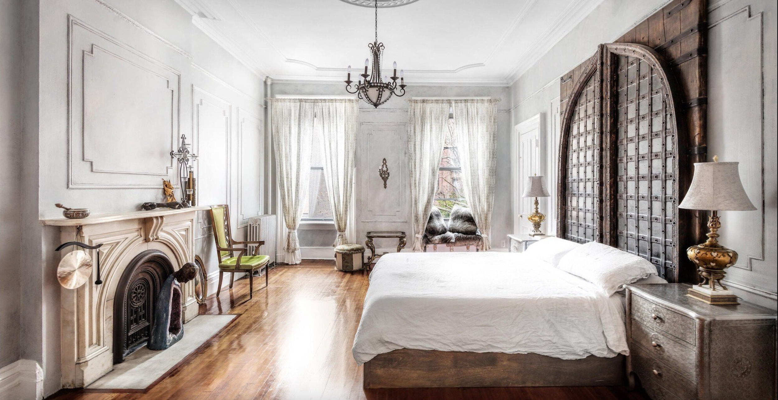 Vintage style bedroom with white walls, hardwood floors, a fireplace, and elegant furniture including a bed with a large wooden headboard, bedside lamps, and a sitting area with chairs and curtains.