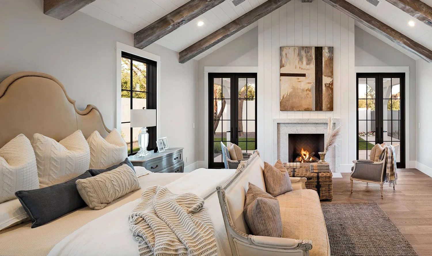A cozy living room with a high ceiling, exposed wooden beams, a fireplace with a painting above, large windows and black-framed glass doors, a sofa with pillows, a beige armchair, and a rug.