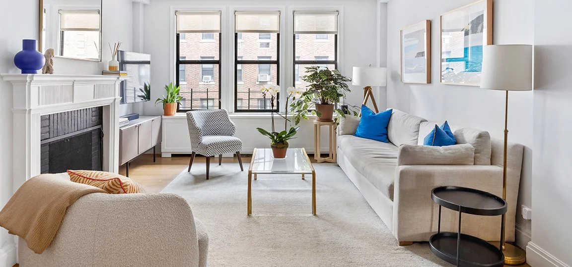 Bright living room with white walls, large windows, beige sofa with blue pillows, a white armchair with a beige blanket, a glass coffee table with a potted plant, and framed artwork on the wall.