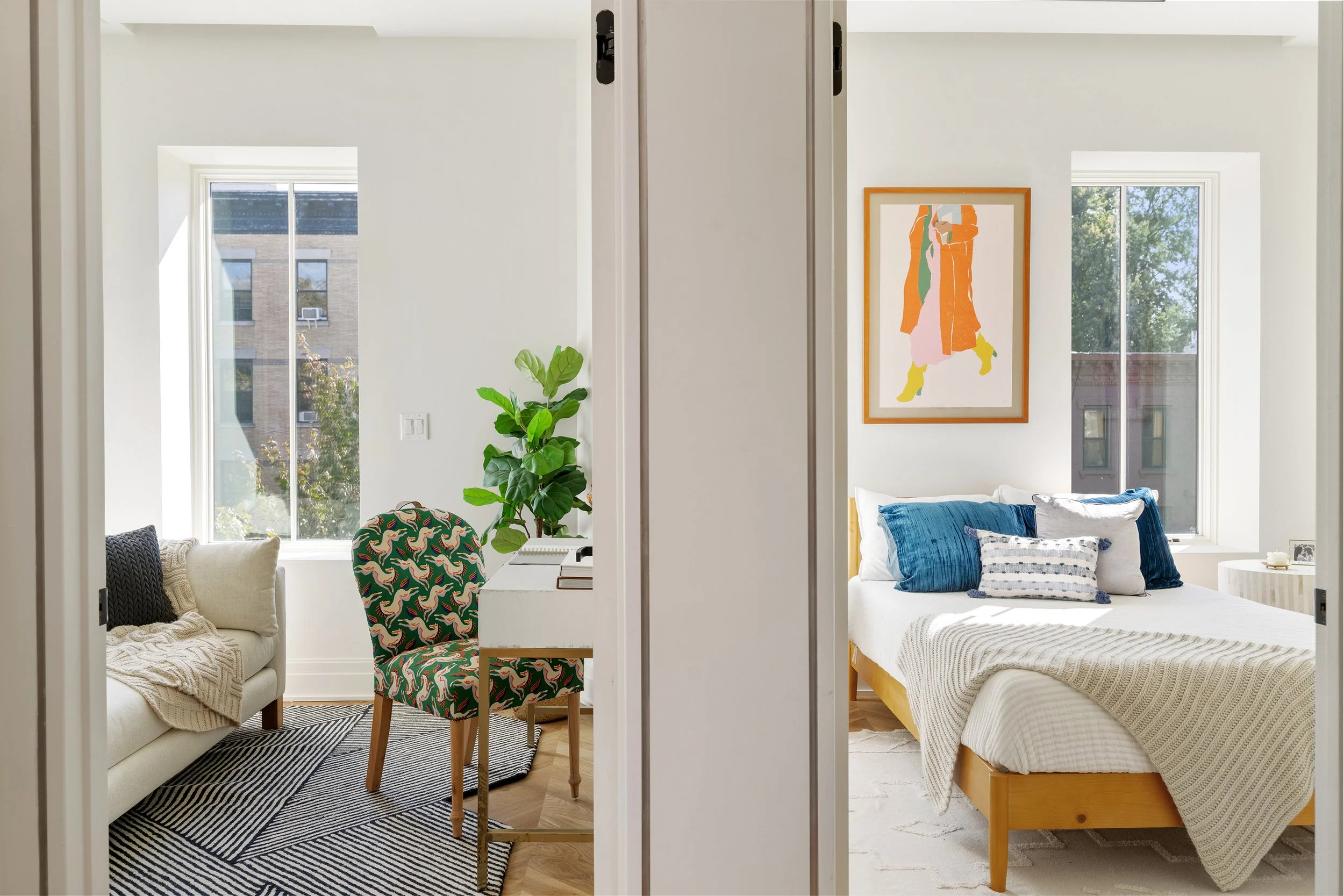 Split view of a modern apartment interior showing a living room on the left with a white couch, a patterned chair, a green plant, and windows; and a bedroom on the right with a bed, blue pillows, framed artwork, and windows with views of trees and bu
