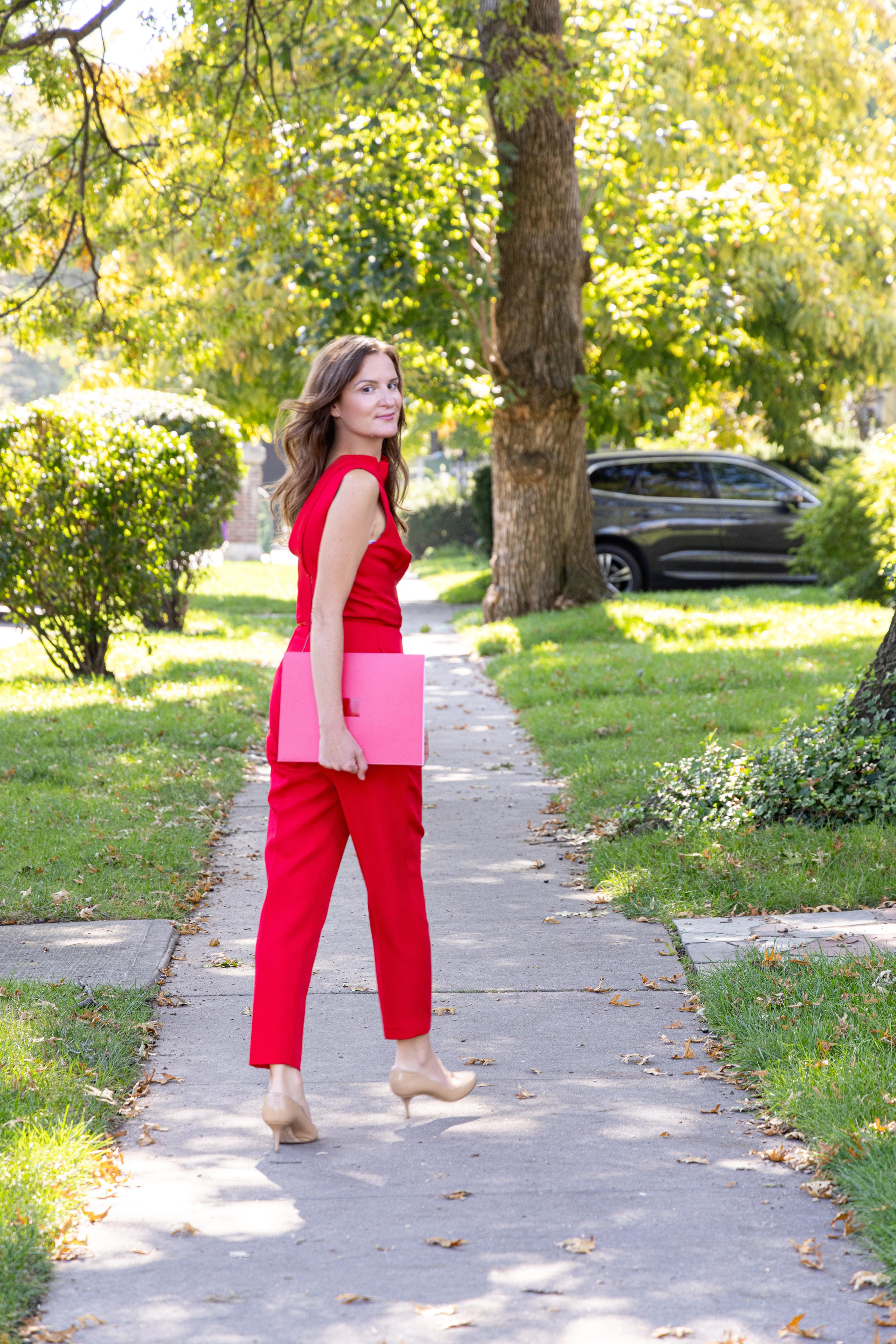 Woman in a red sleeveless jumpsuit holding a pink folder and standing on a sidewalk in a green park.