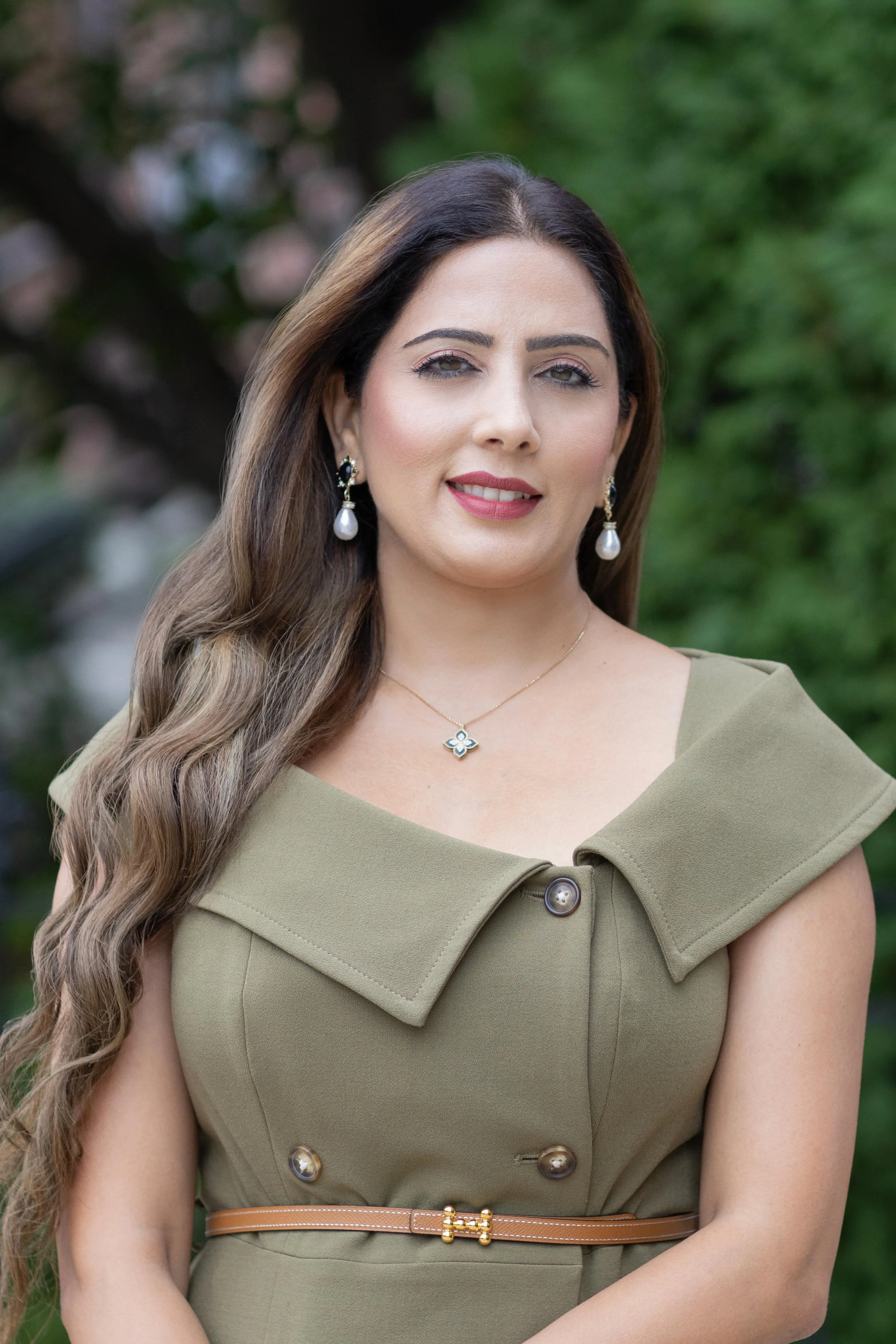 A woman with long, wavy brown hair and light makeup wearing pearl earrings, a gold necklace, and an olive green dress with large collar details and buttons, standing outdoors with blurred green foliage in the background.