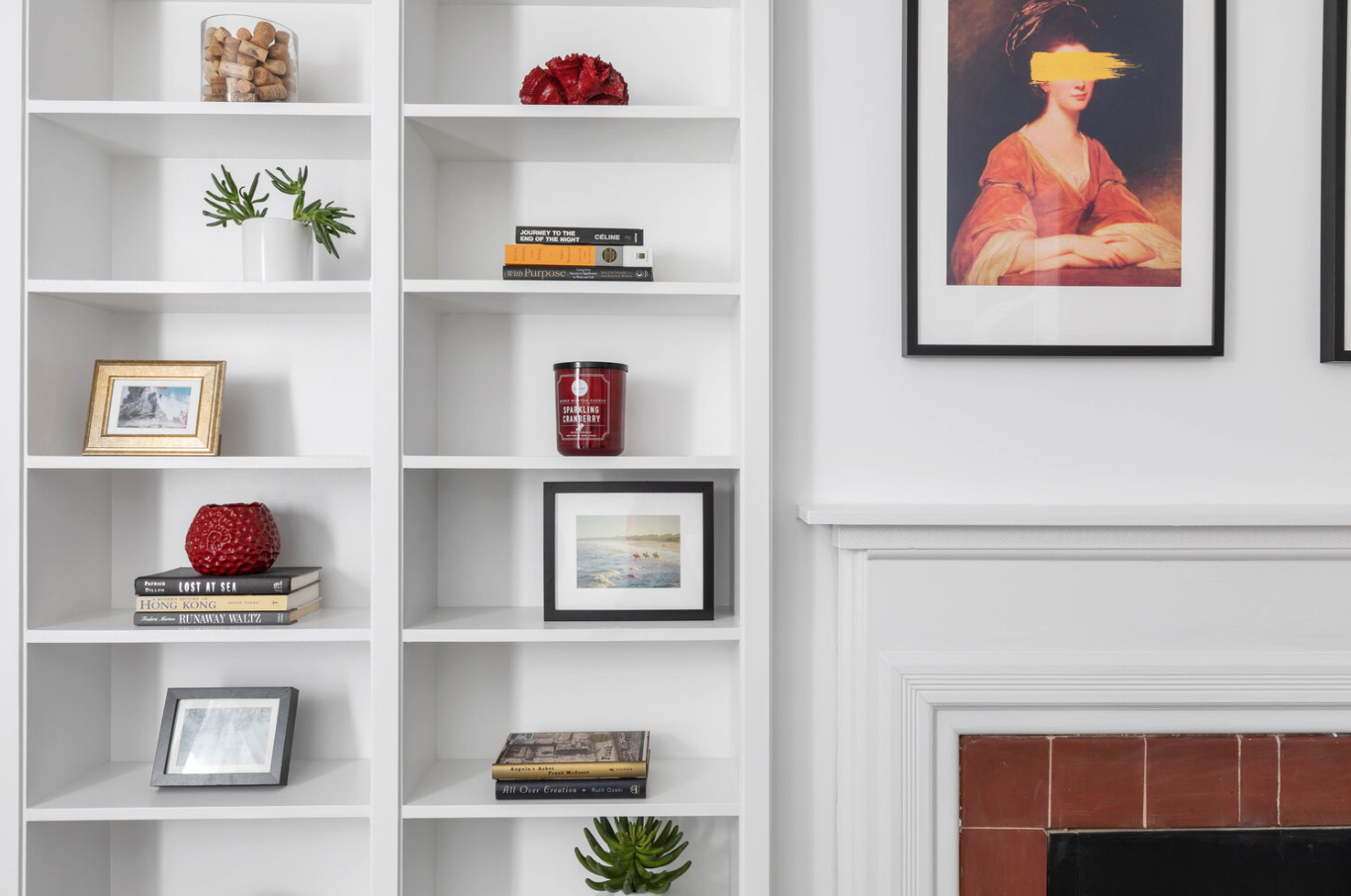 White bookshelf with decorative items, books, and framed photos in front of a white wall with art.