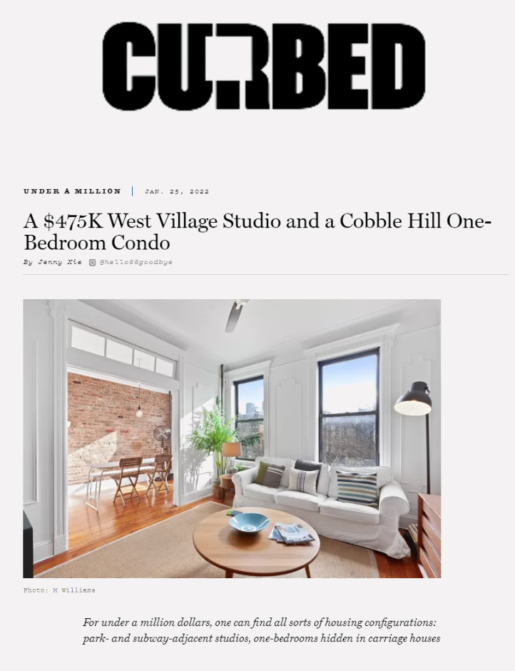 Bright living room in a modern condo with white walls, two large windows, a white sofa with striped cushions, a round wooden coffee table with bowls and magazines, a potted plant, a standing lamp, and a view into a dining area with a brick wall and wooden chairs.