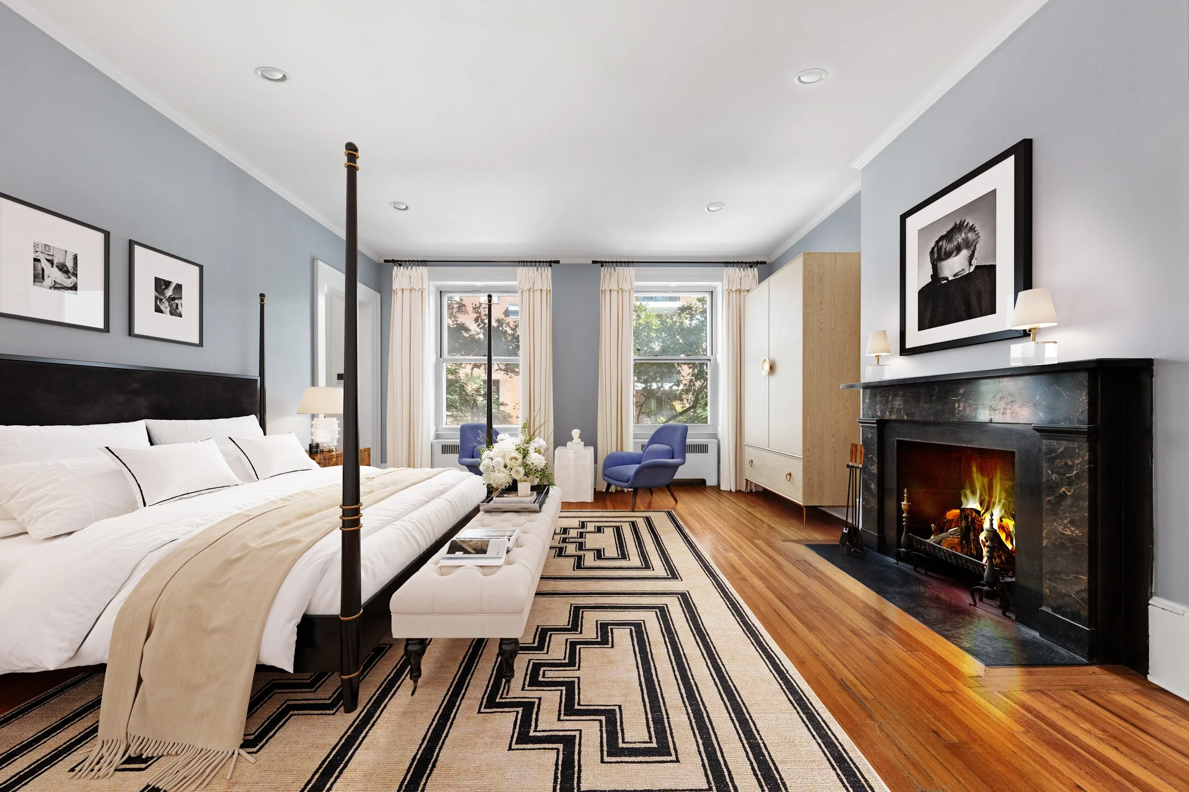 Living room with blue walls, four-poster bed, black and white striped rug, white and beige bedding, two blue armchairs, fireplace with a photograph of a person above it, and wooden floors.
