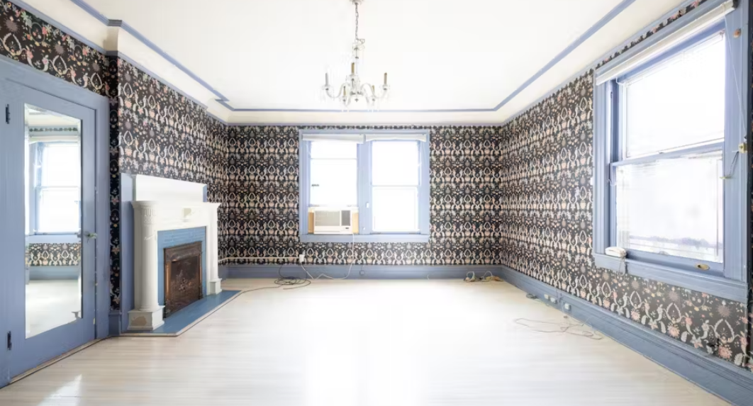 Empty living room with floral wallpaper, hardwood floors, a chandelier, and windows with air conditioning unit.