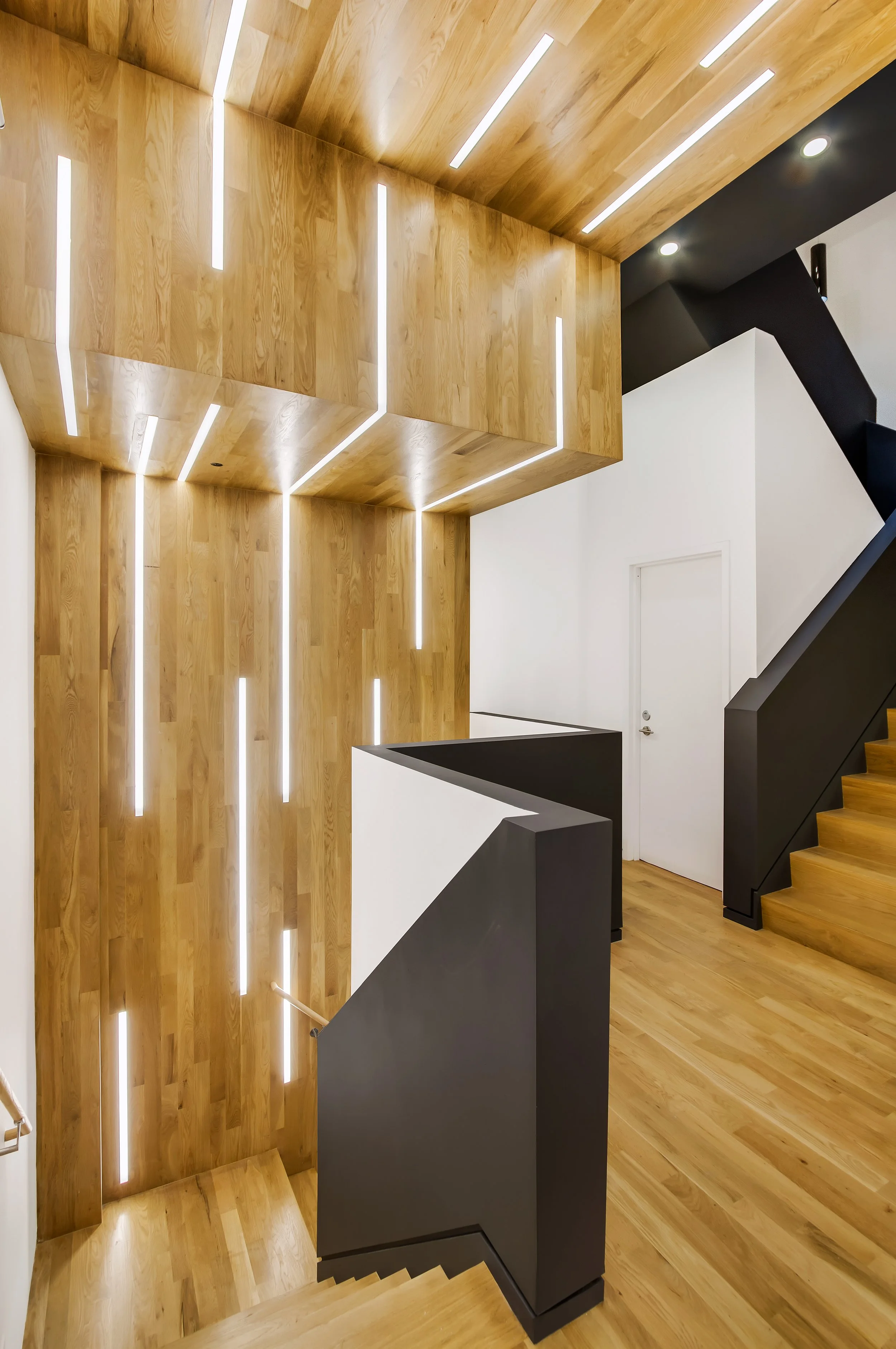 Modern staircase with wooden steps and black and white walls, featuring decorative vertical LED lights on a wood-paneled wall.